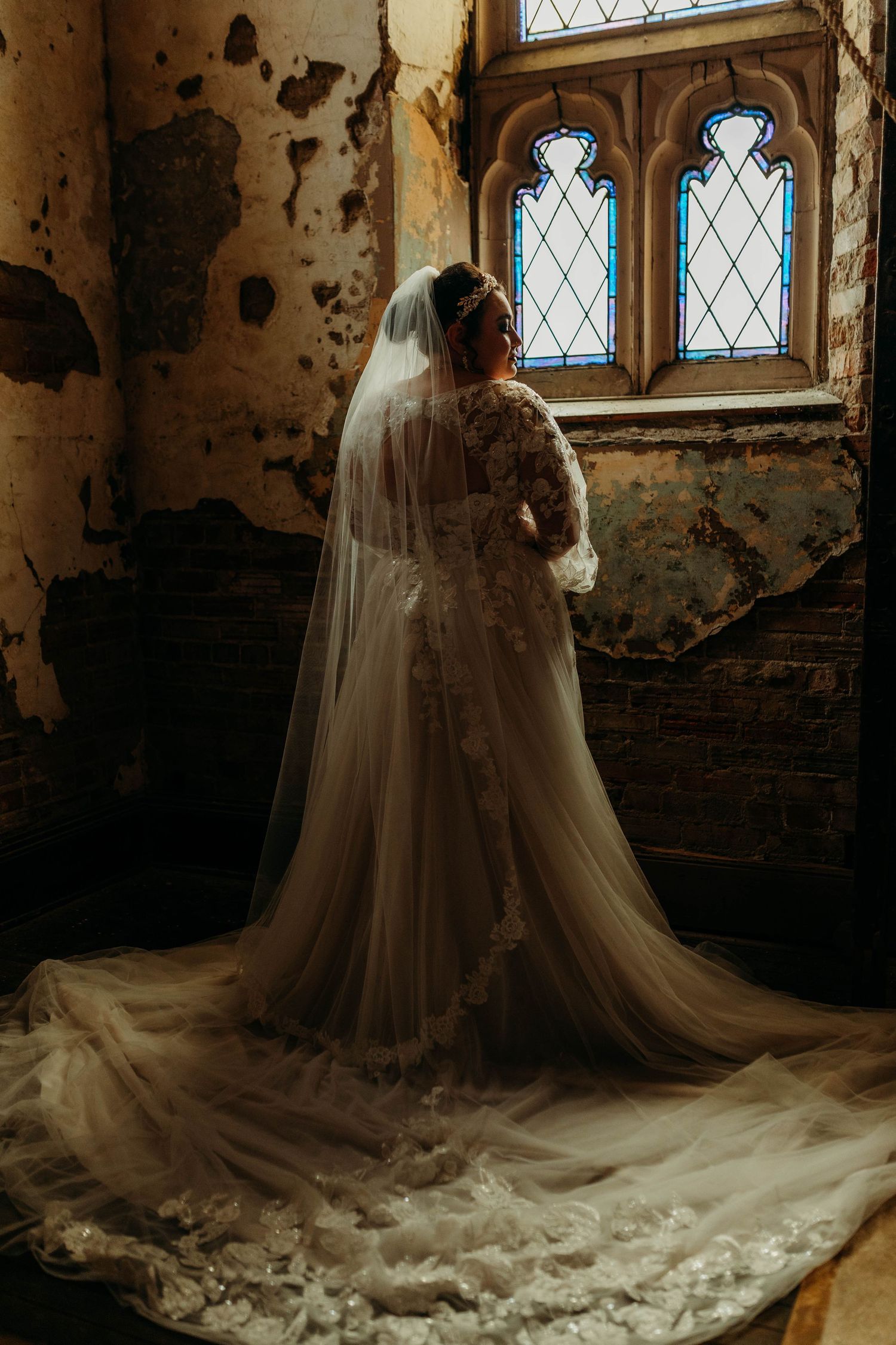 A dramatic wedding dress with a flowing train photographed in an old building with distressed walls and gothic windows.
