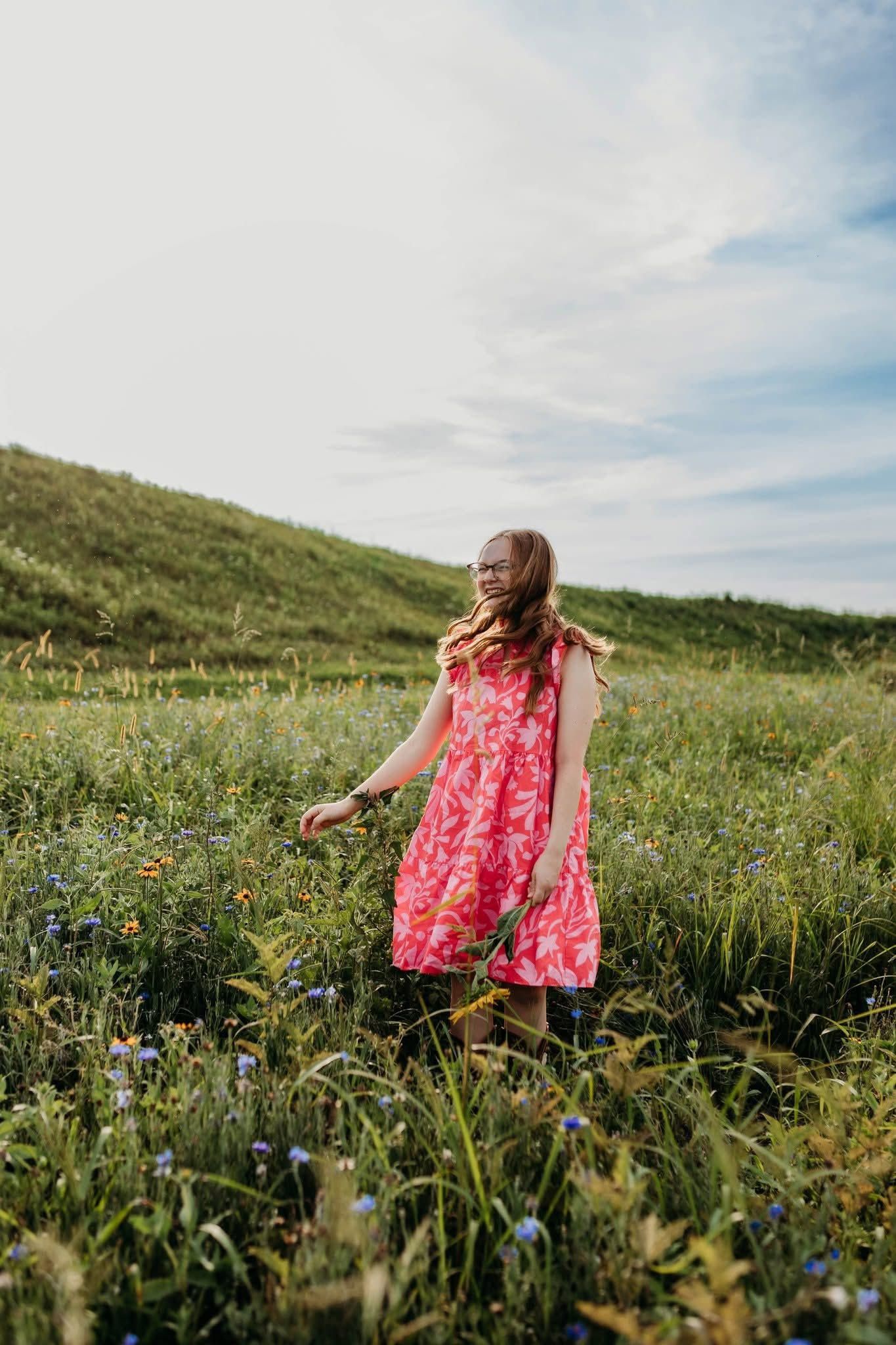 A young girl in a pink floral dress walks through a lush green meadow with wildflowers under a partly cloudy sky.
