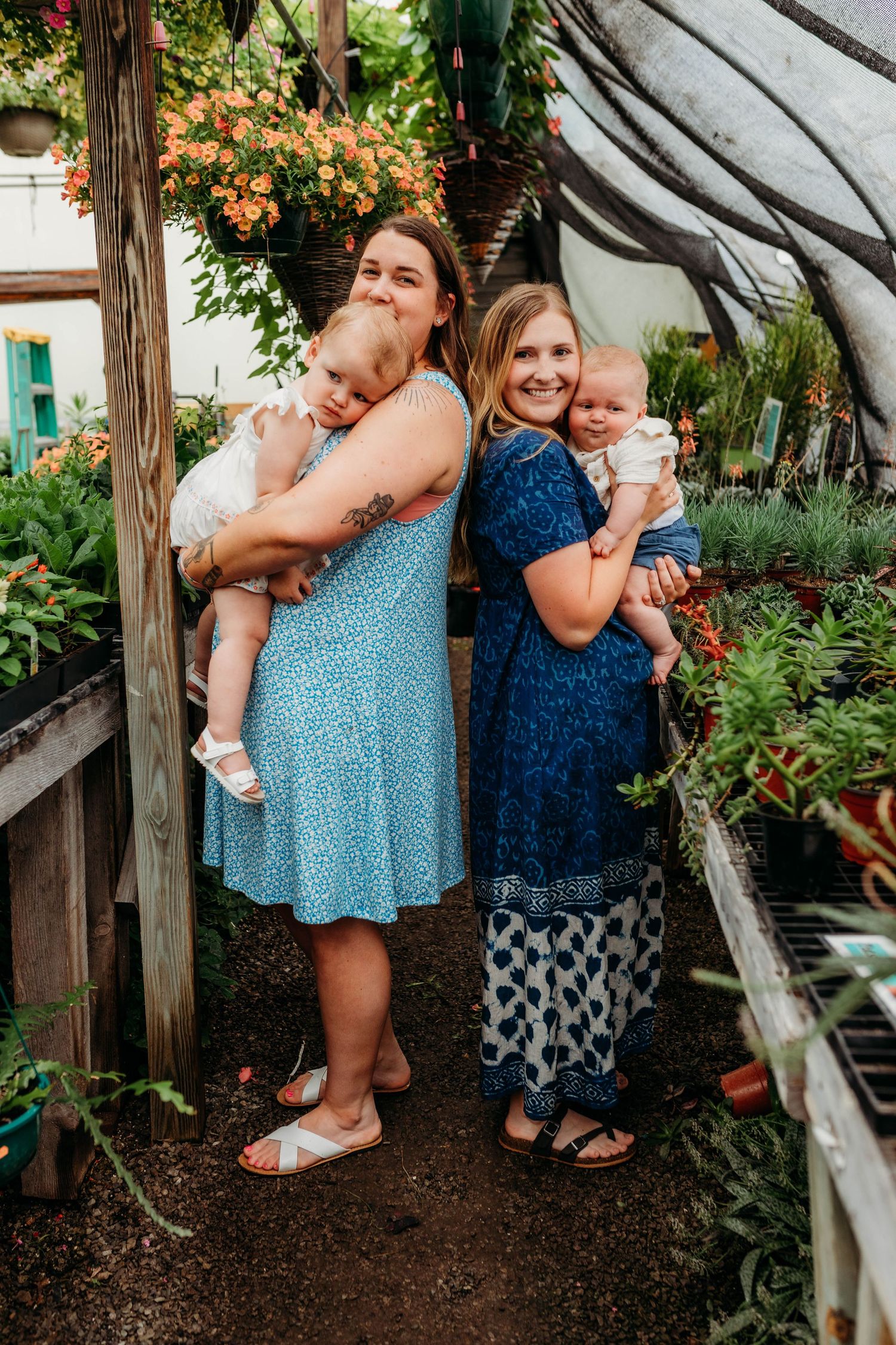 Two adults holding babies while posing together in a greenhouse with flowering plants.