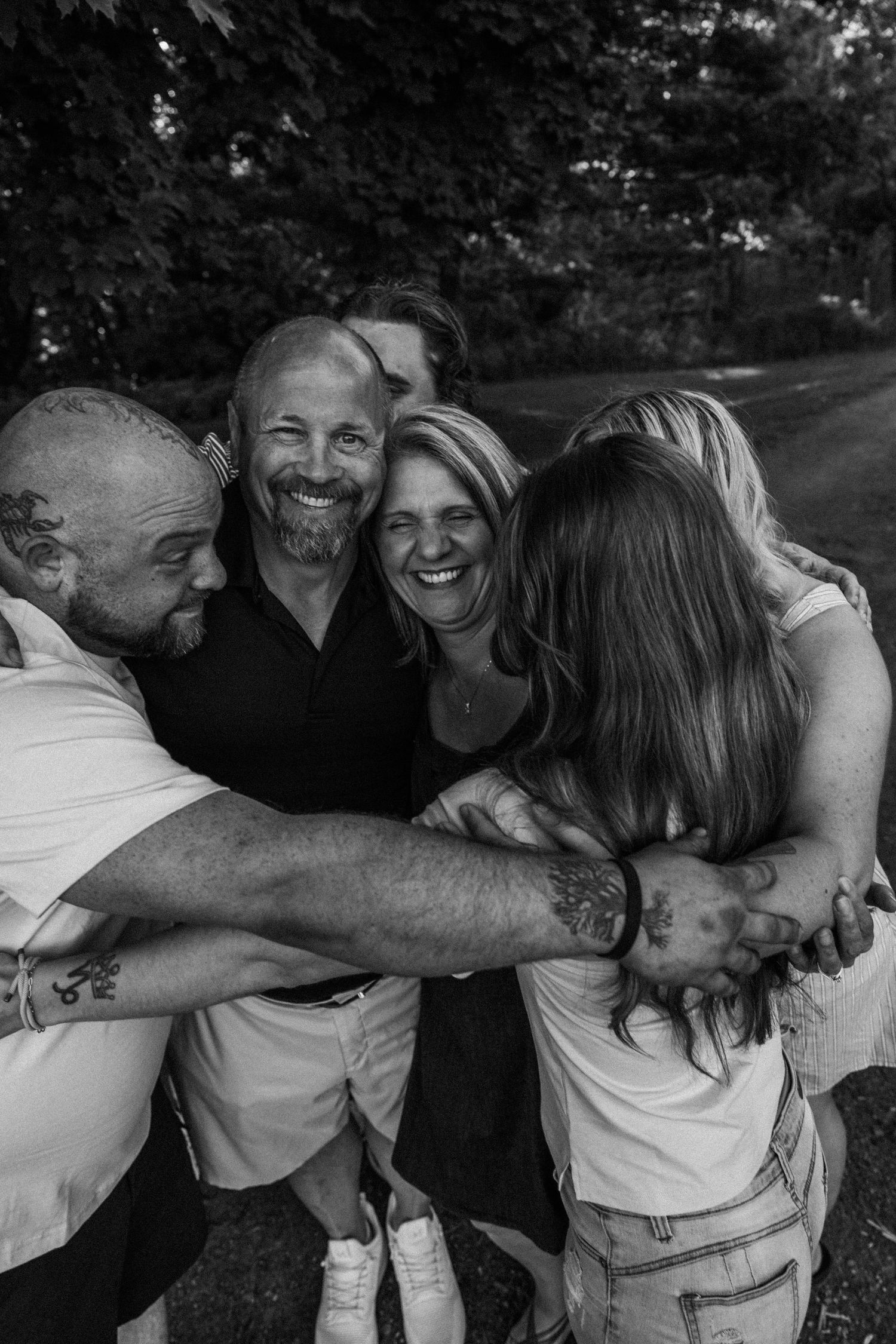 Black and white photo of a joyful family group hug outdoors, smiling adults embracing tightly among lush trees.