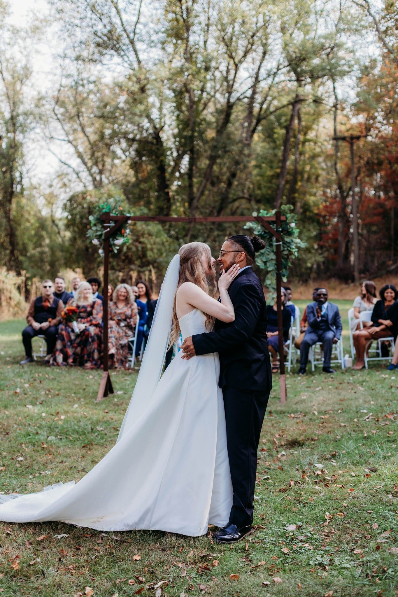 Wedding ceremony kiss under floral arch with guests seated in woodland setting.