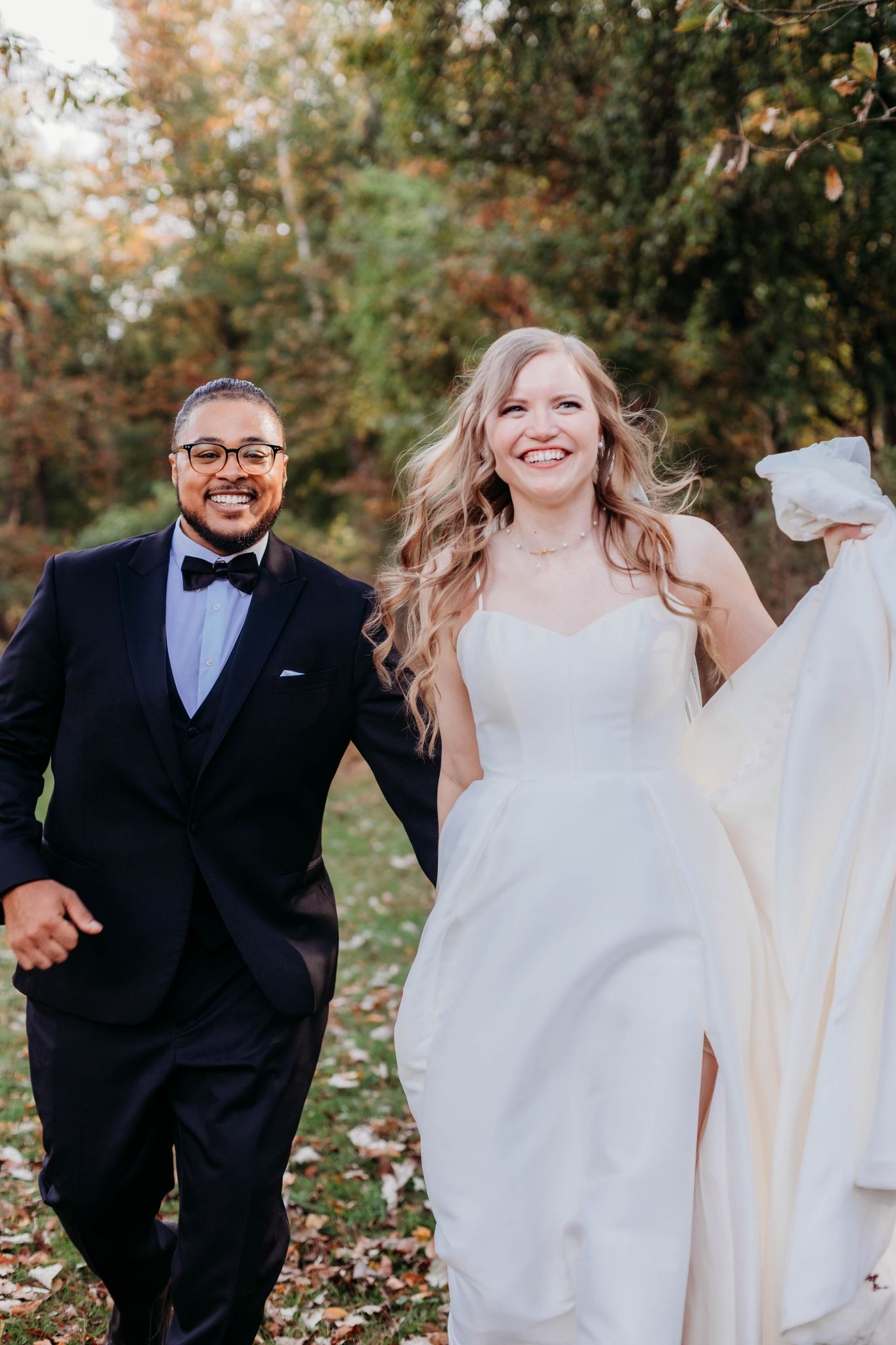 Joyful wedding couple playing and running through autumn leaves in formal attire.