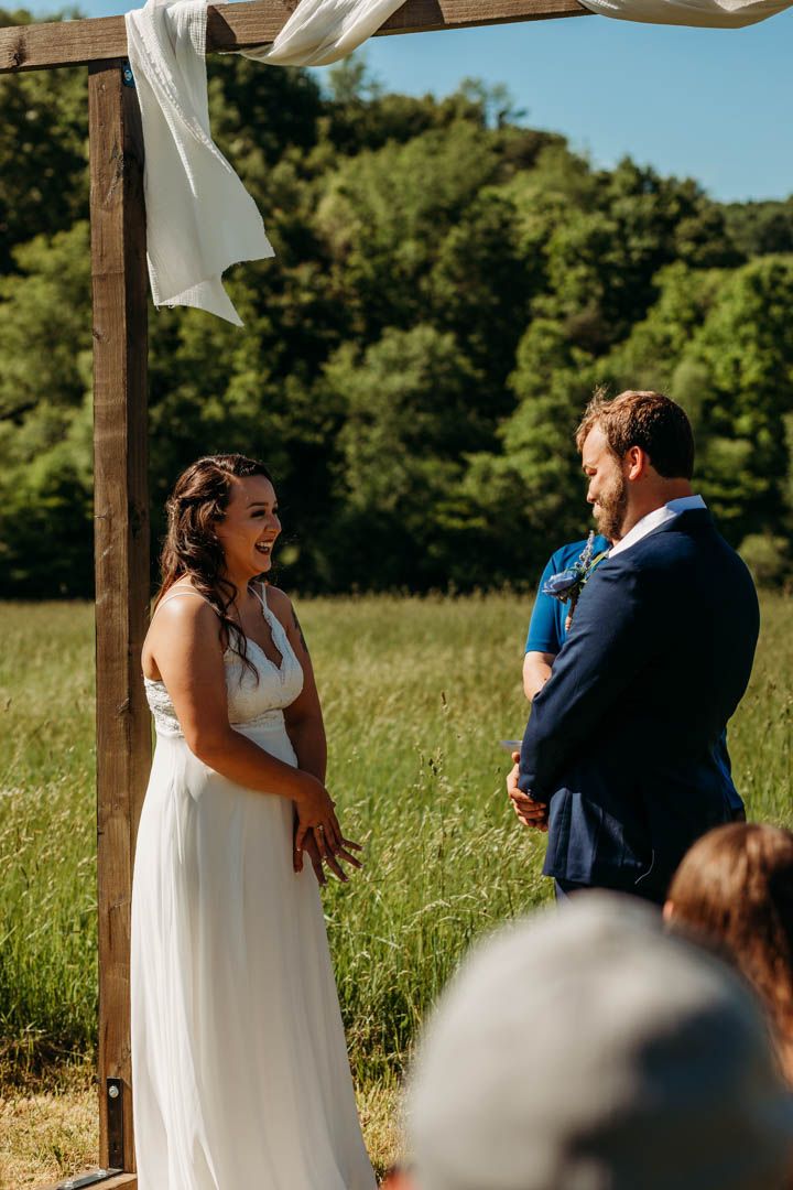 Outdoor wedding ceremony under wooden arch with white fabric in sunny field.
