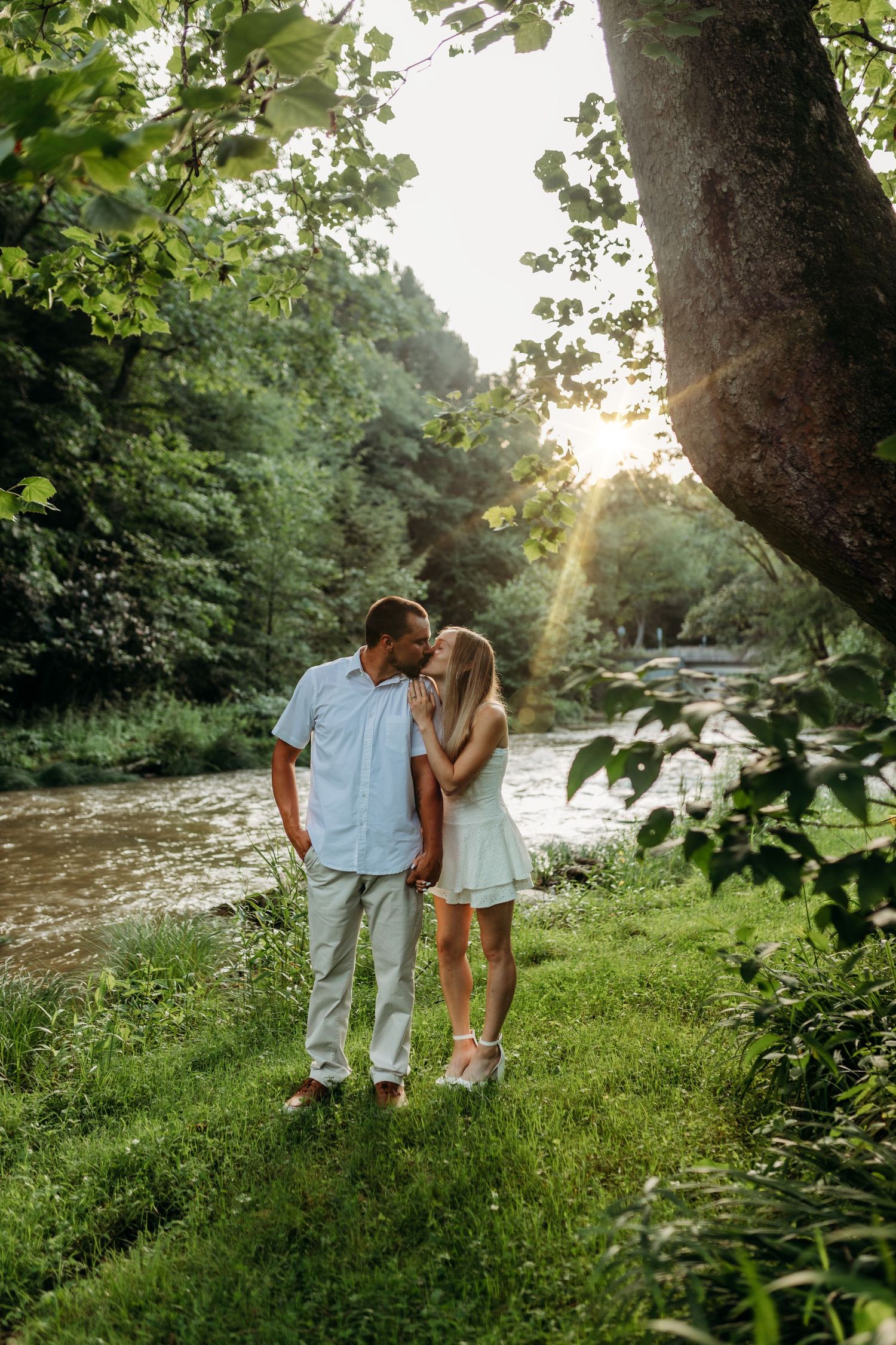 Romantic couple standing by river at sunset surrounded by lush greenery.