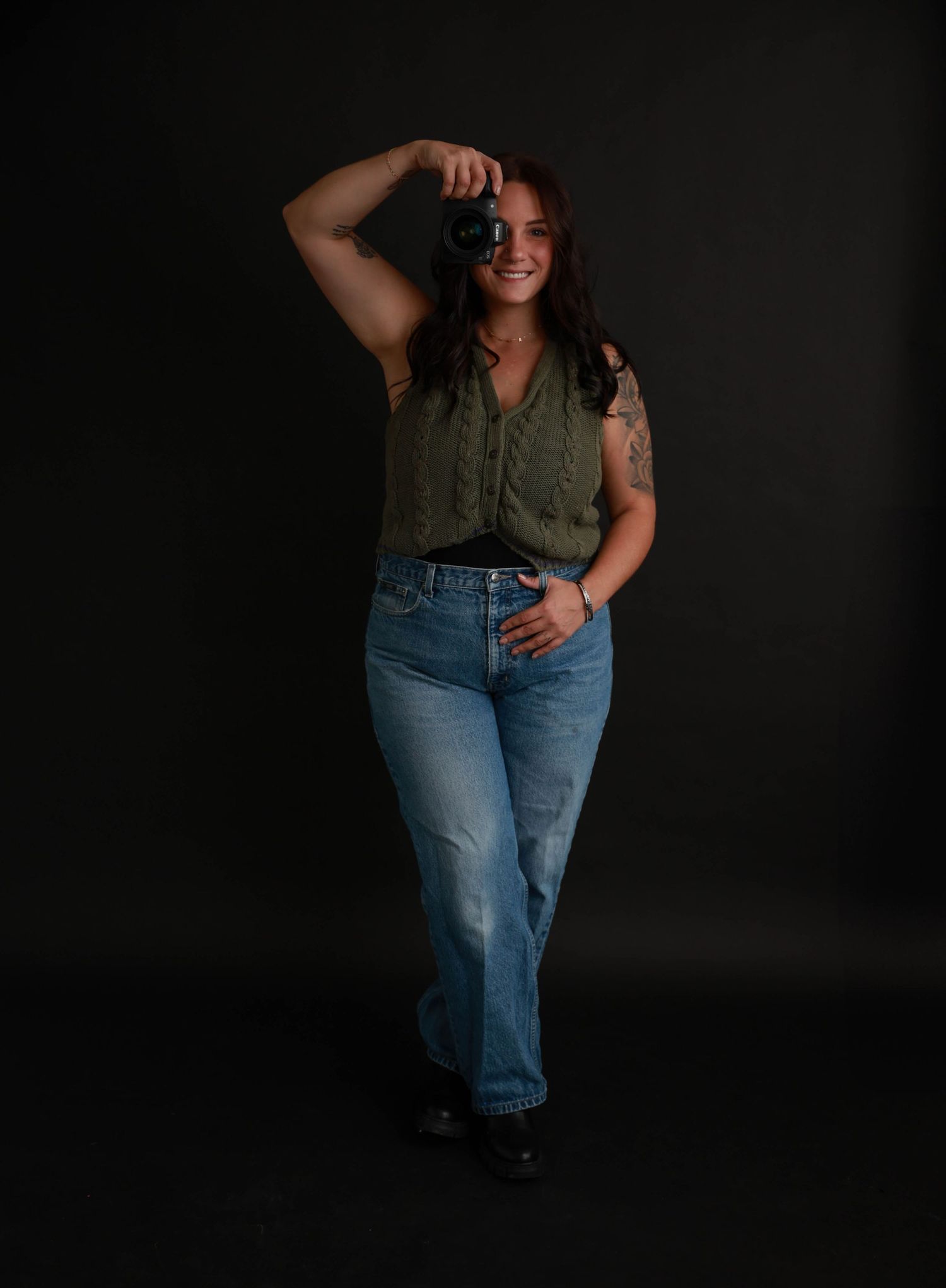 Professional photographer in olive top and jeans poses with camera against dark backdrop.