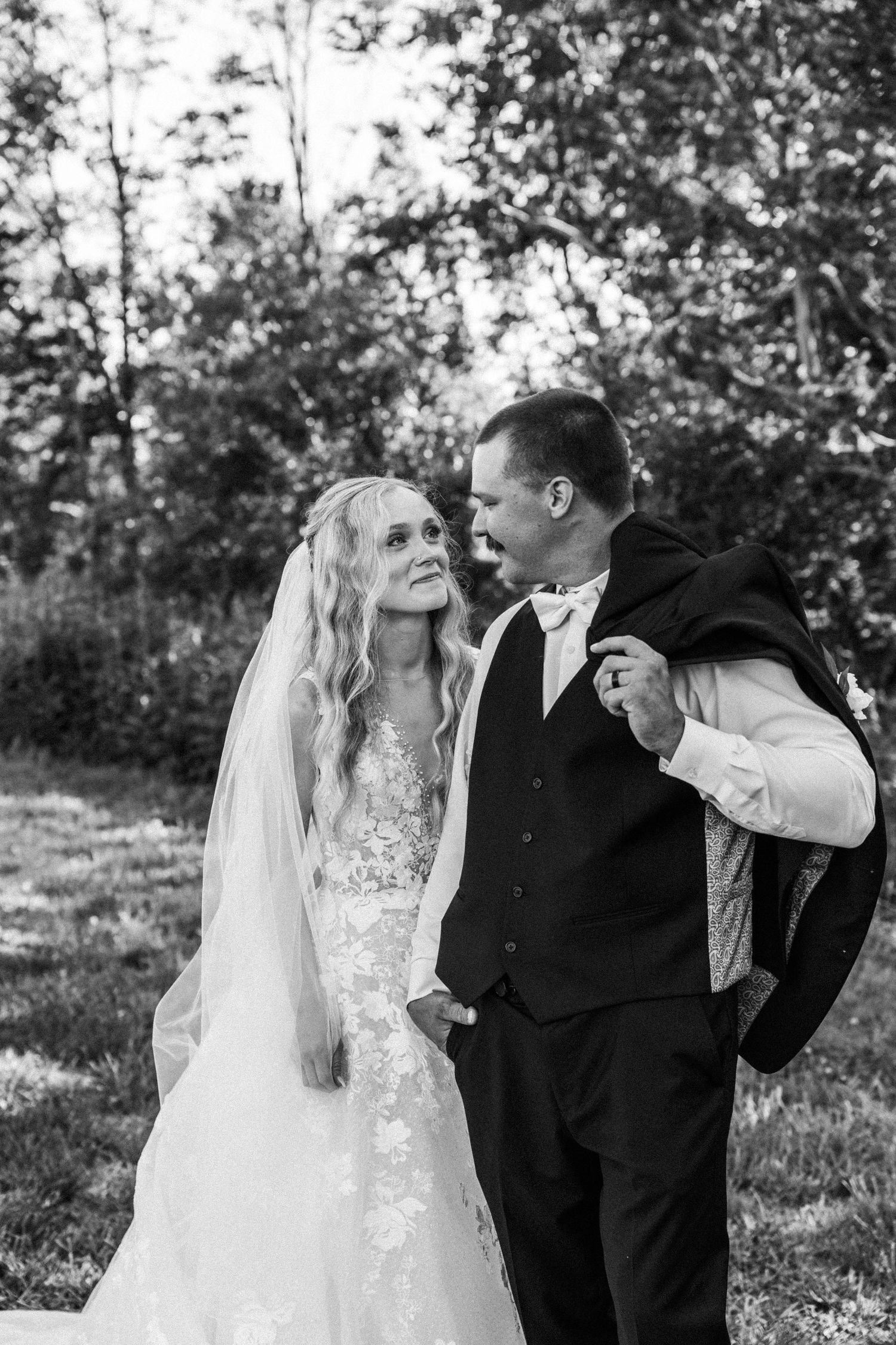 Black and white photo of a bride and groom sharing a tender moment outdoors, surrounded by lush green trees.