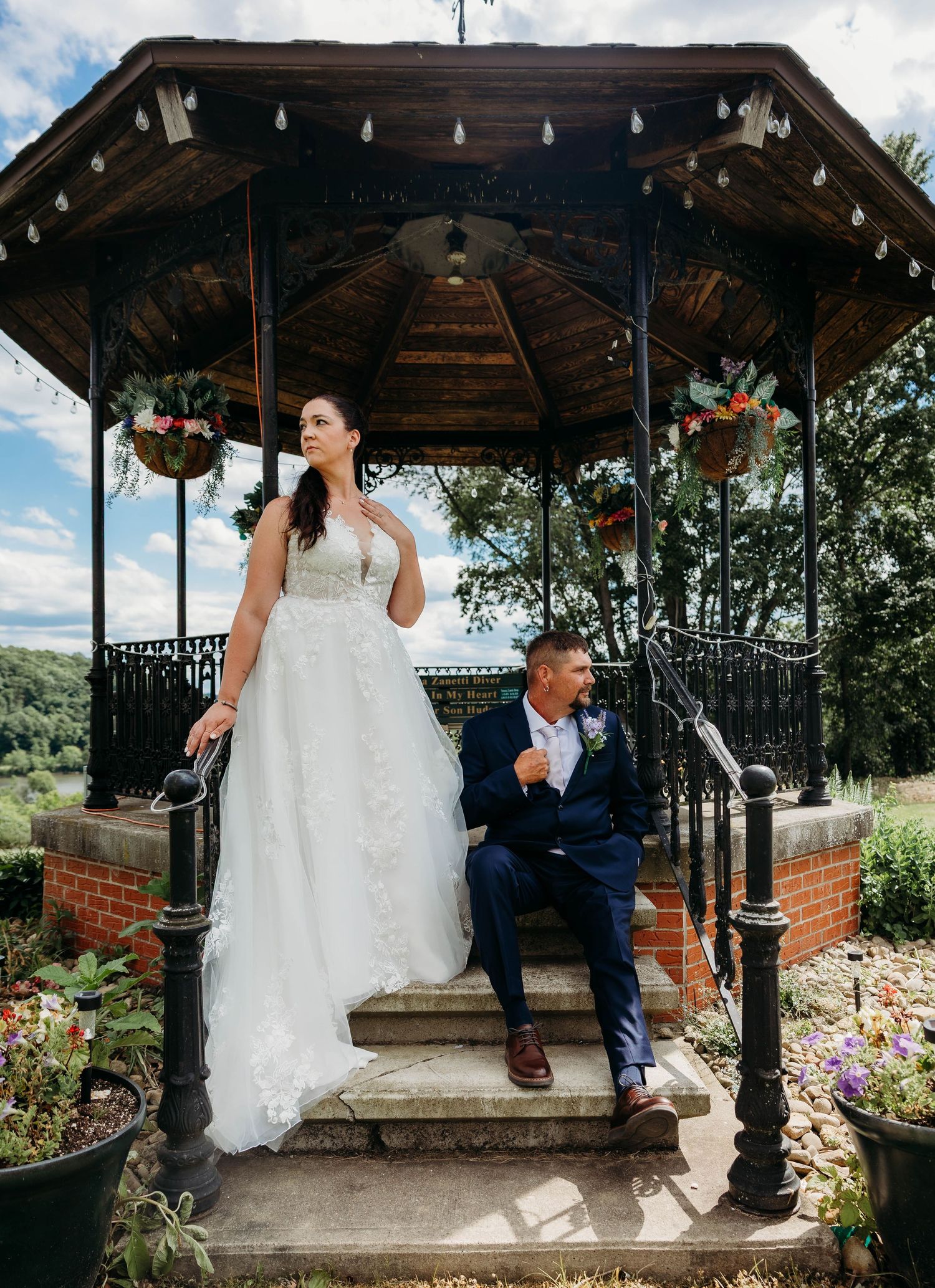 Bride in white gown and groom in navy suit pose at a decorated gazebo surrounded by lush gardens on their wedding day.