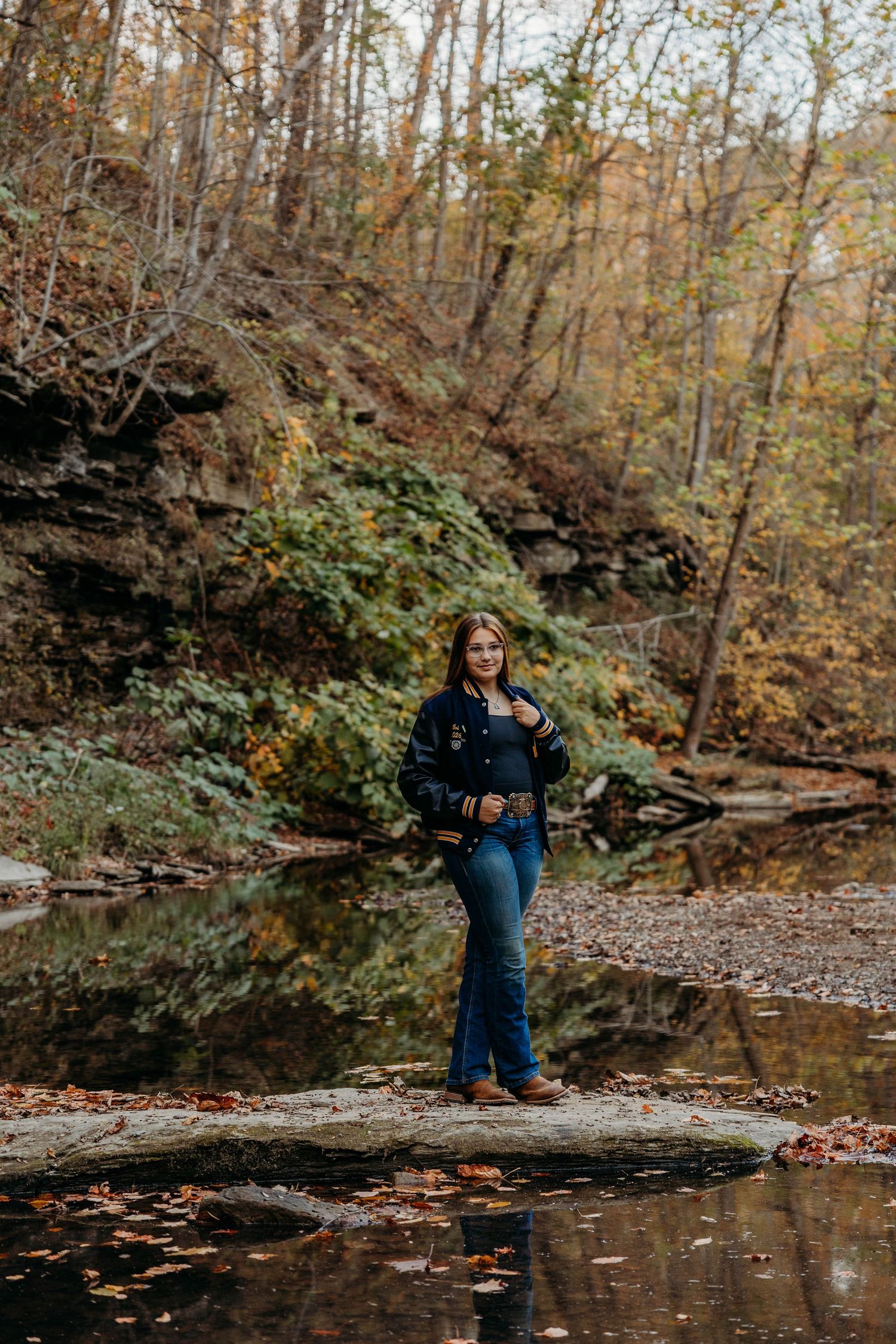 Young woman in dark jacket and jeans standing on rocks by a reflective creek in an autumn forest.