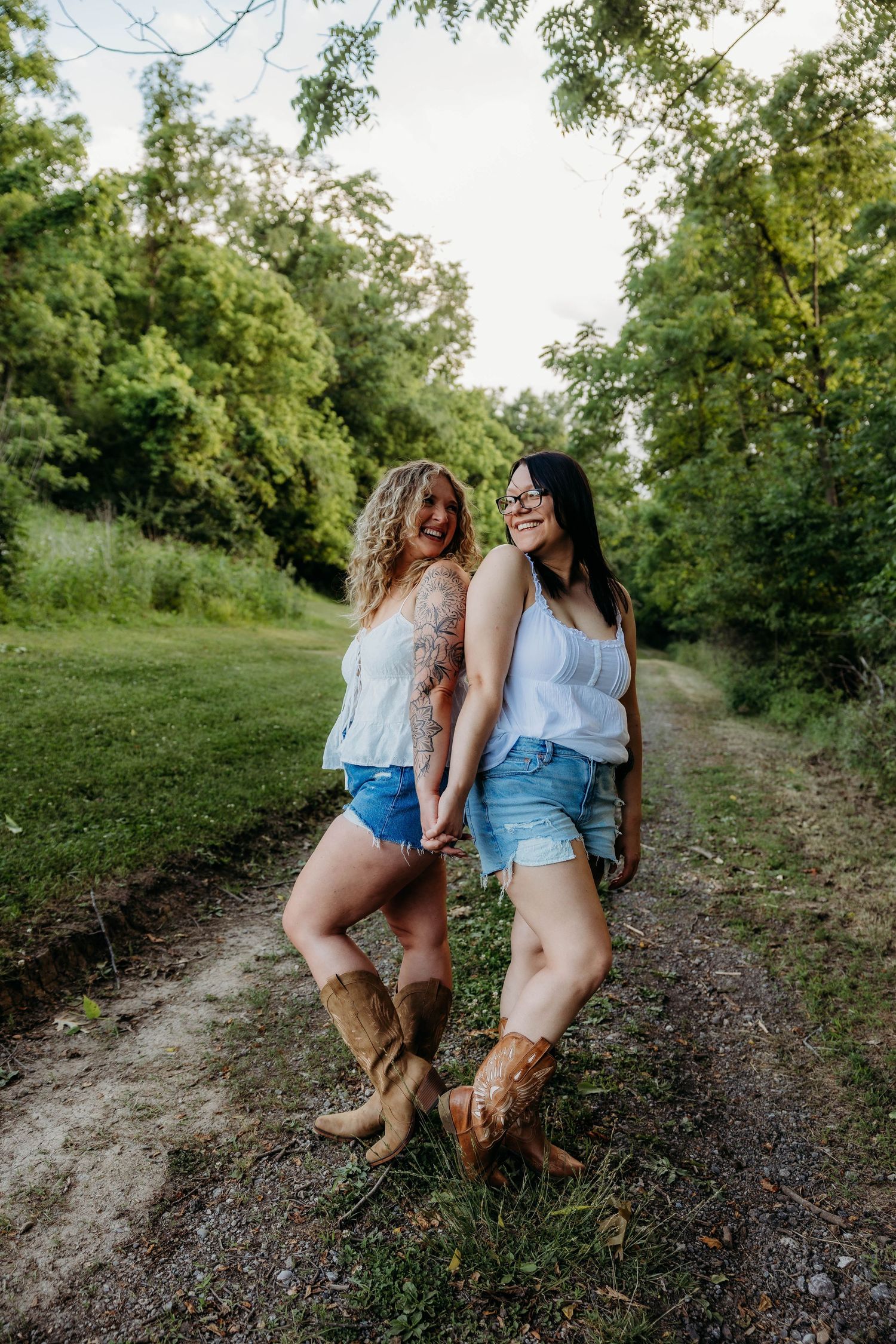 Two women in white tops, denim shorts, and cowboy boots hold hands on a rural path surrounded by green trees.