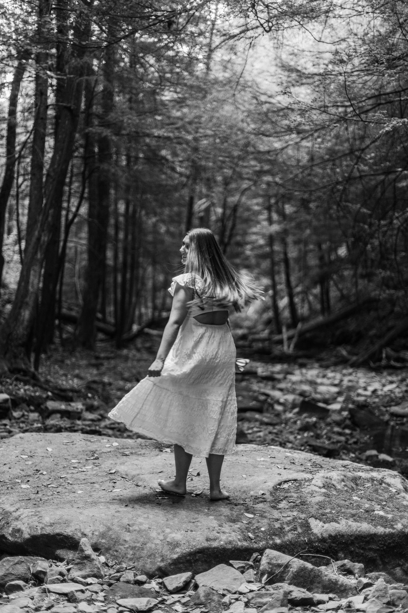 A dreamy black and white photo of a woman in a dress standing on a rock in a misty forest.