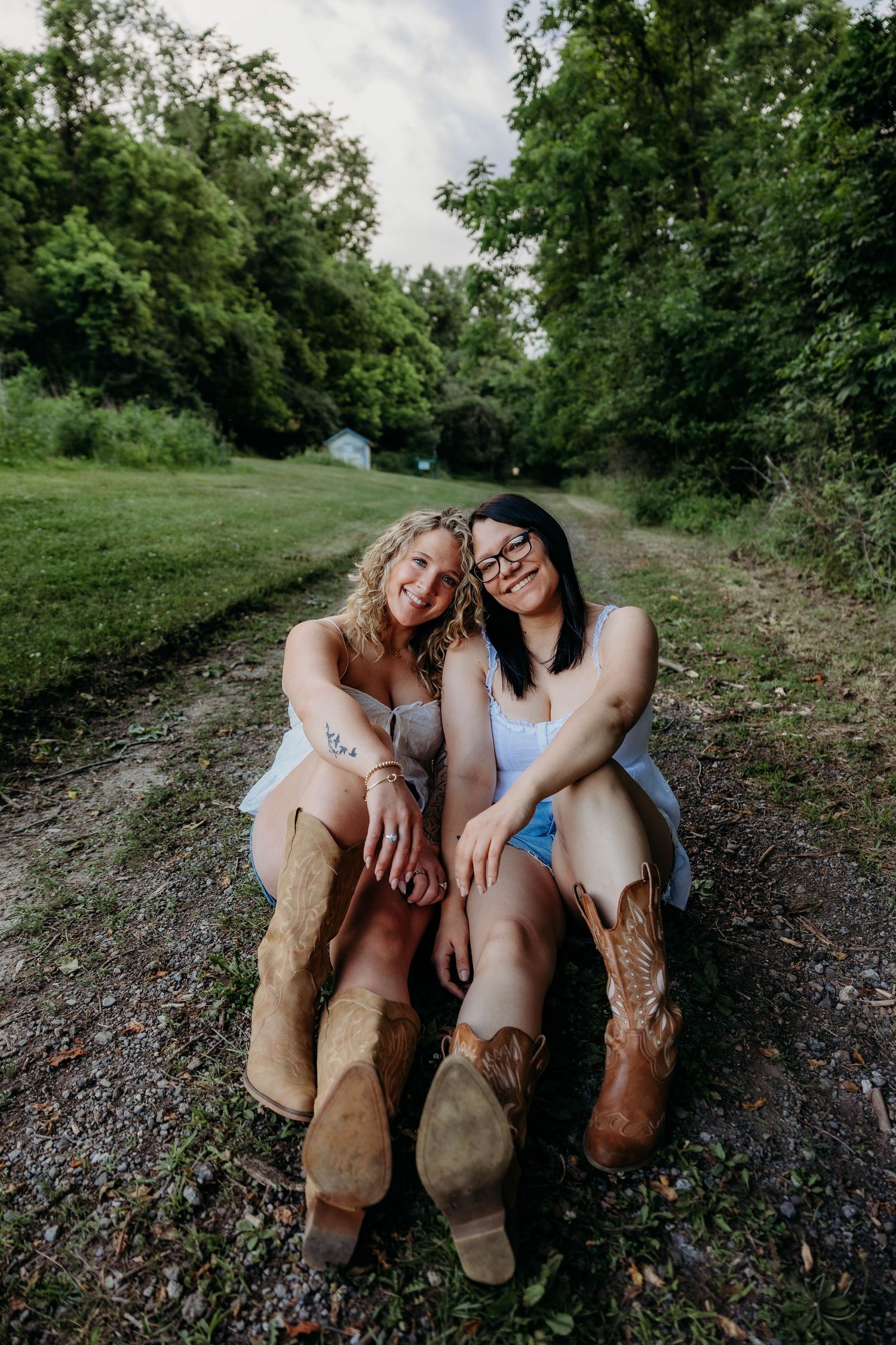 Two smiling women in summer dresses and cowboy boots sit together on a rural dirt path surrounded by lush green trees.
