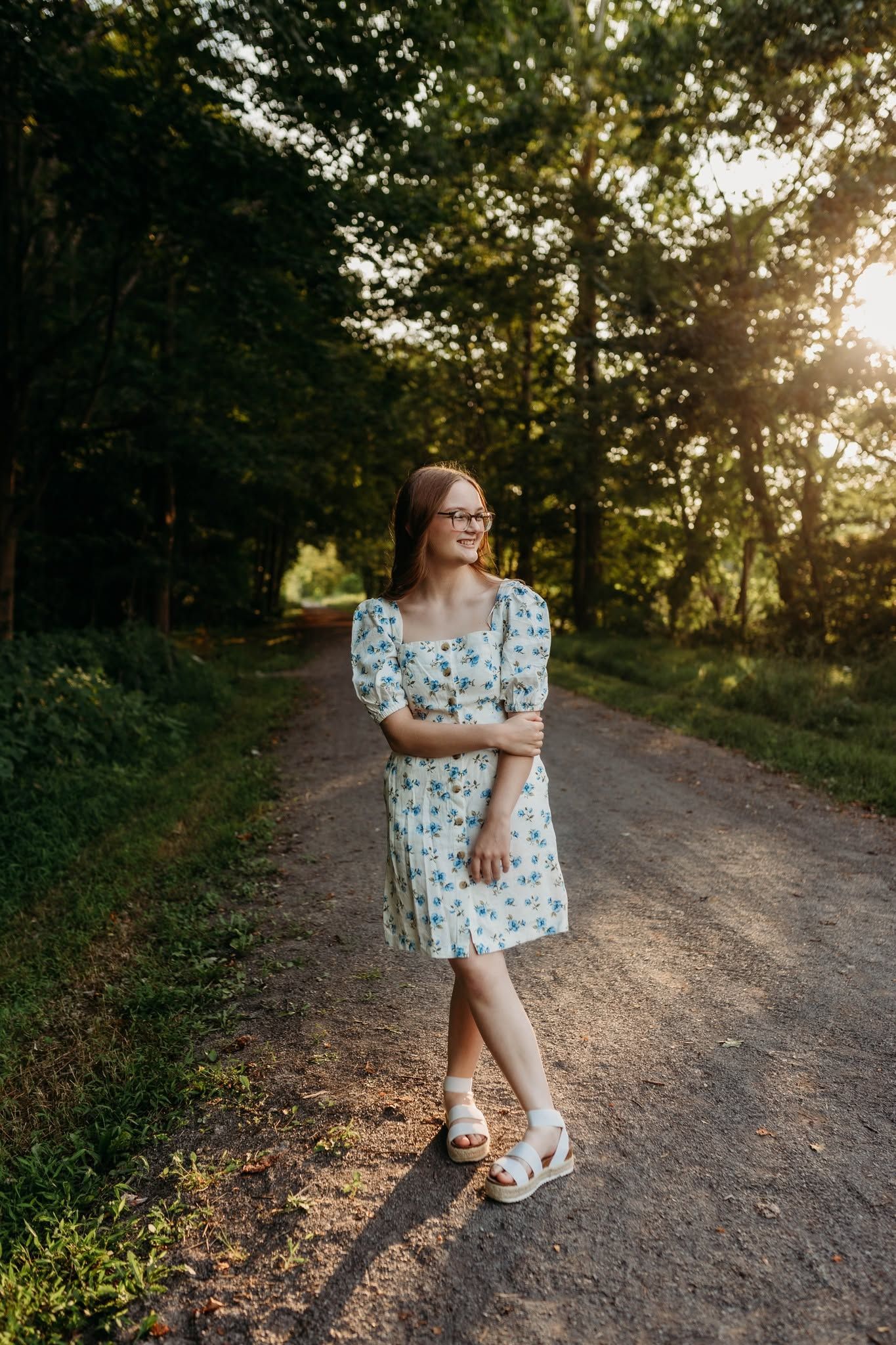 Young woman in floral dress and glasses standing on a sunlit forest path during golden hour.
