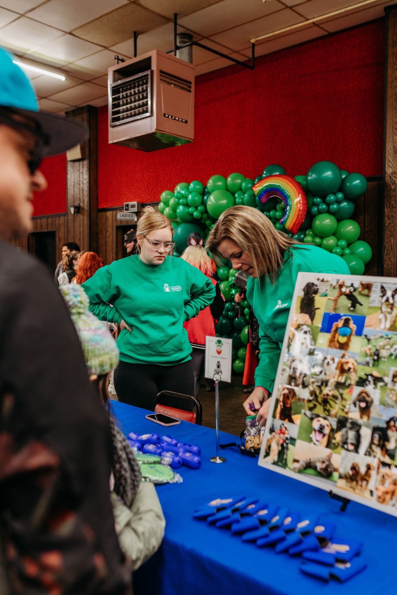 Event staff in green shirts arrange items on a blue table with green balloon decorations in the background.