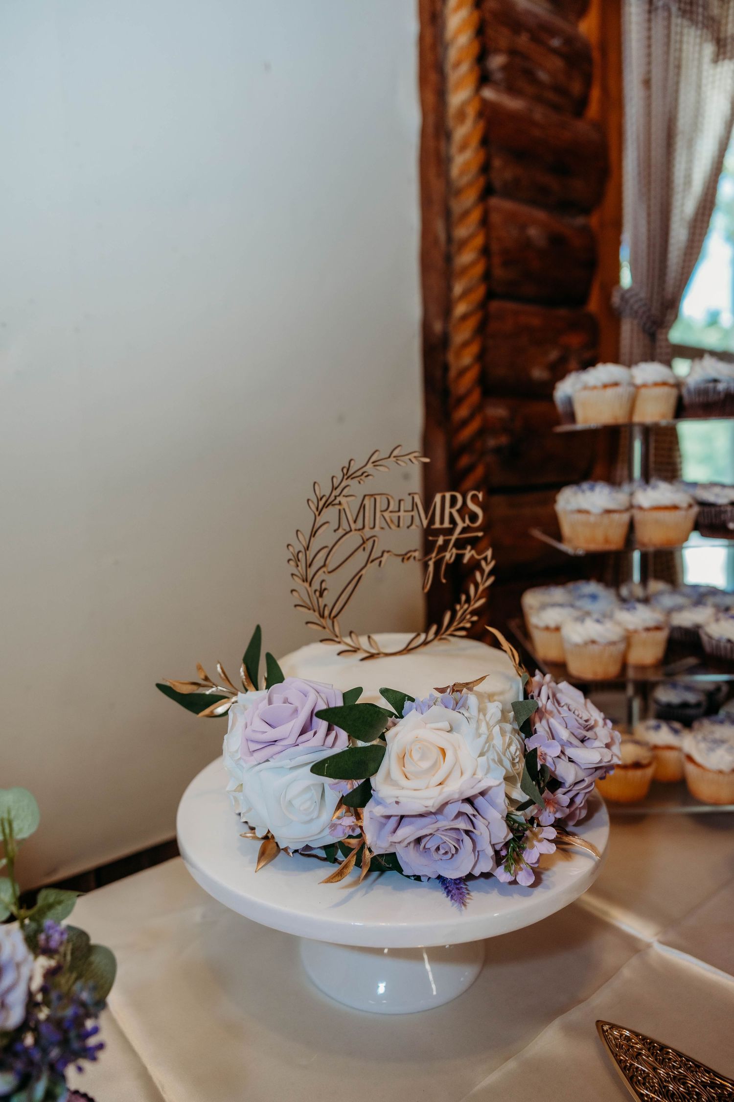 Elegant one-tier wedding cake with purple and white roses, Mr & Mrs topper on white stand, with cupcake tower in background.