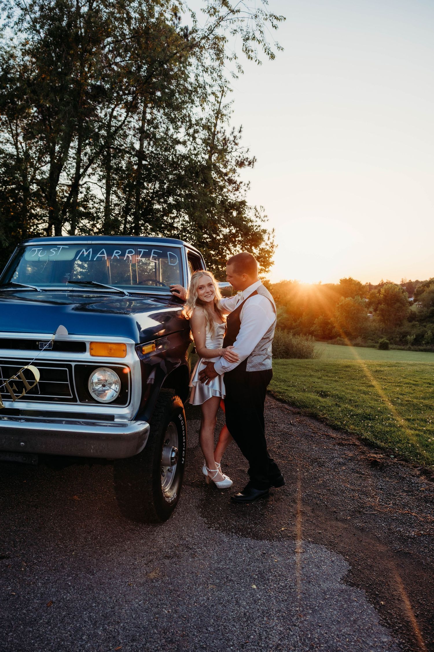 Newlywed couple embracing beside a vintage blue truck at golden hour sunset, with Just Married sign.