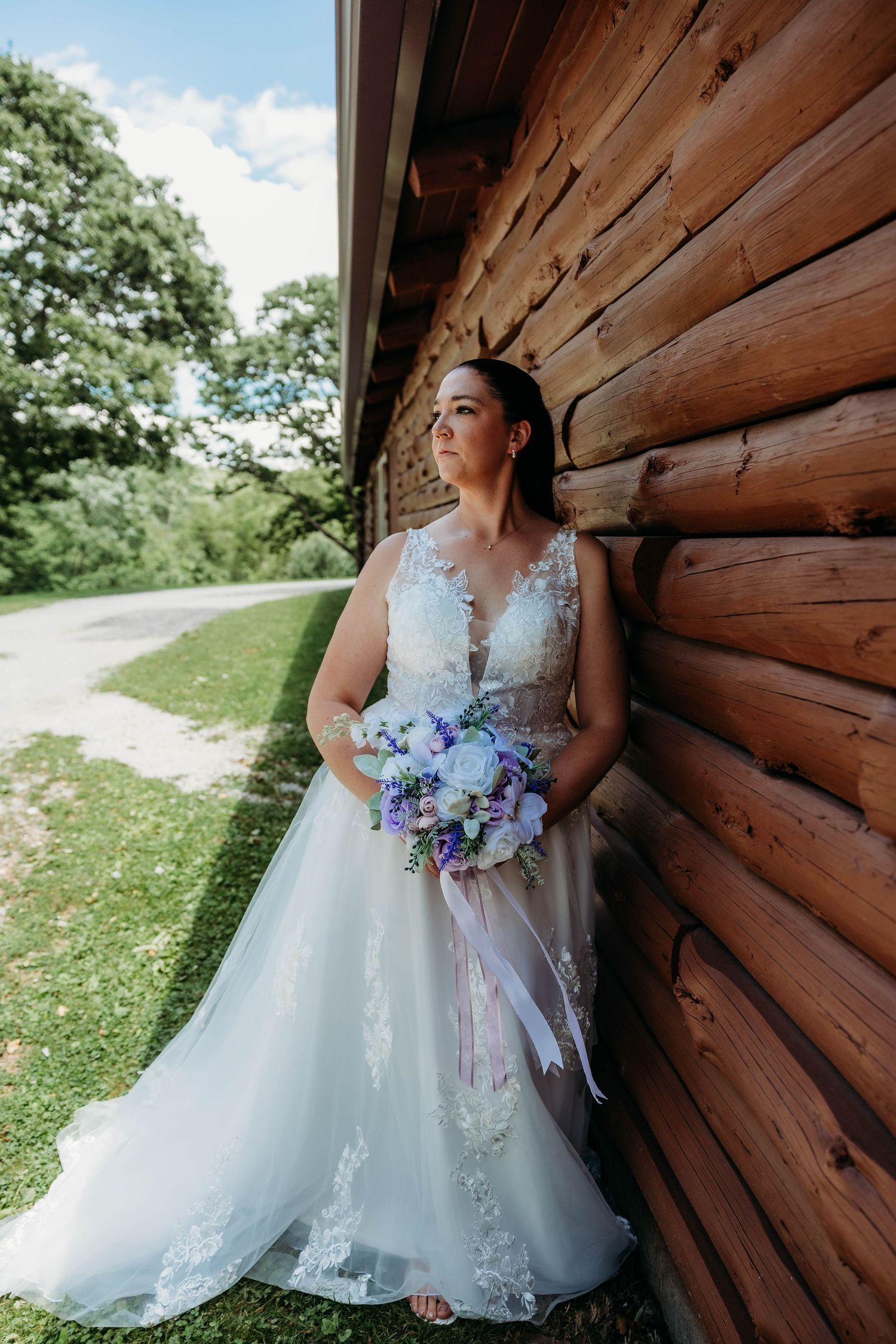 Bride in lace wedding gown holding purple and white bouquet, leaning against rustic log cabin wall outdoors.