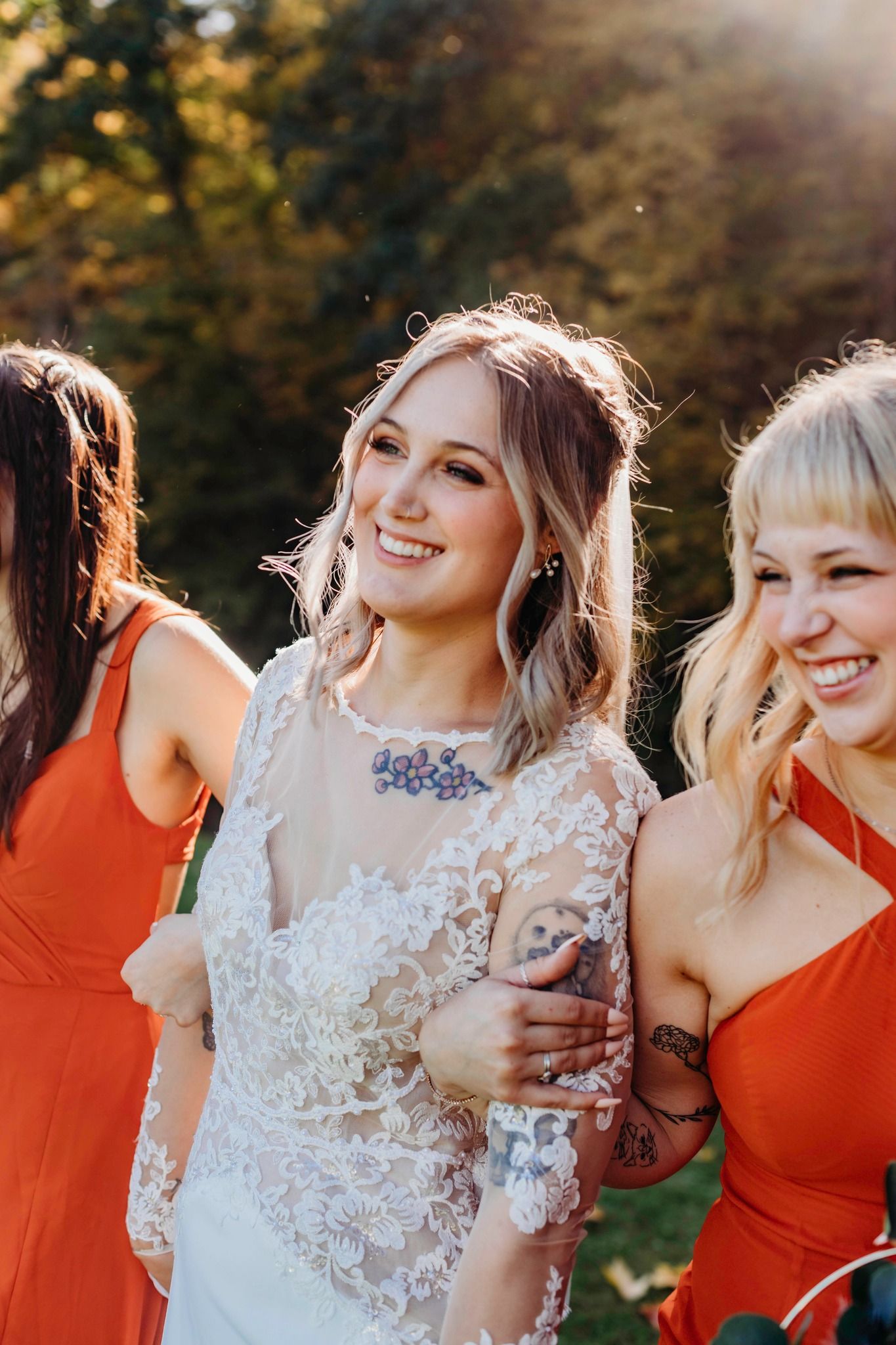 Bridal party in white lace and orange dresses laugh together in autumn sunlight.