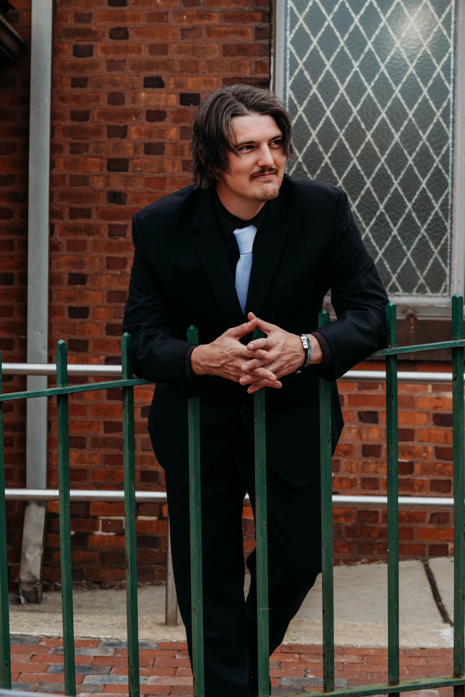 Men in dark suits leaning against a green railing outside a brick building with diamond-paned windows.