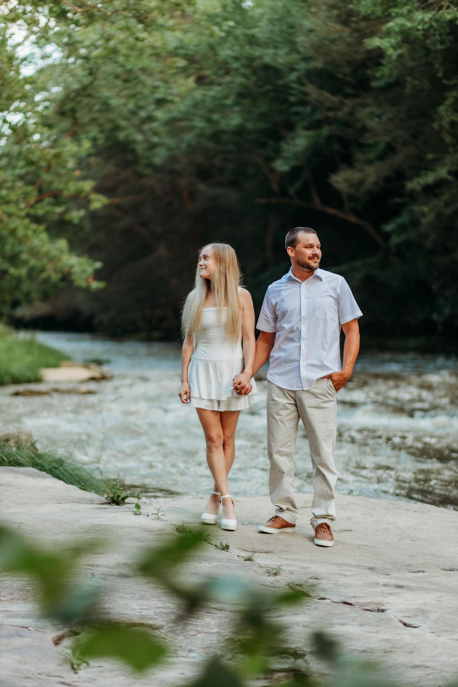 A couple takes a casual stroll along a rocky riverbank on a sunny day.