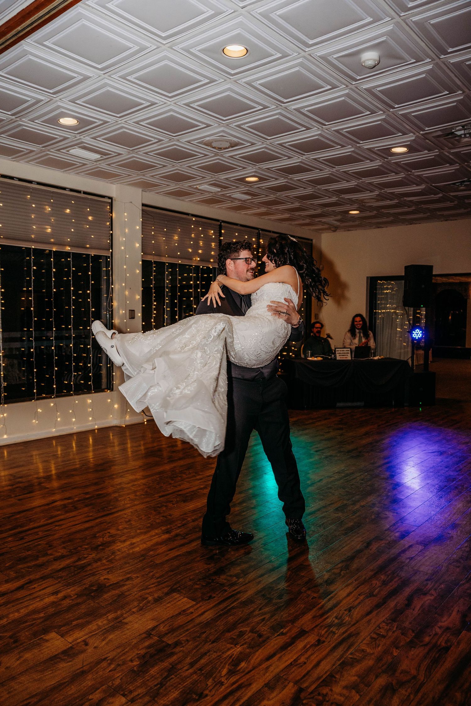 A couple shares their first dance at a wedding reception with twinkling lights and hardwood floors under ornate ceiling.