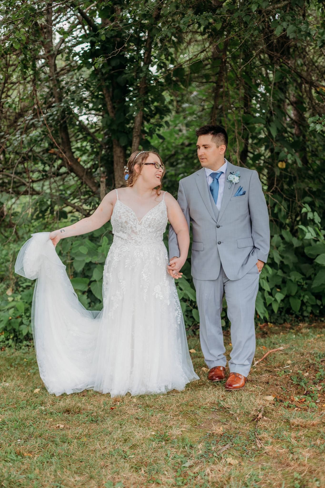 Newlyweds in grey suit and white lace gown pose for romantic photos on grassy hillside.