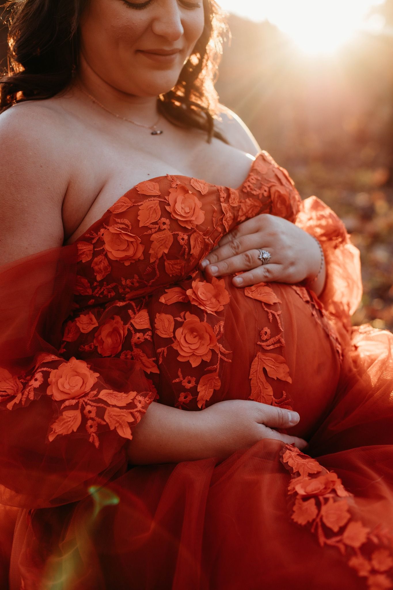 Maternity photo showing pregnant woman in flowing red lace dress against golden sunset light.