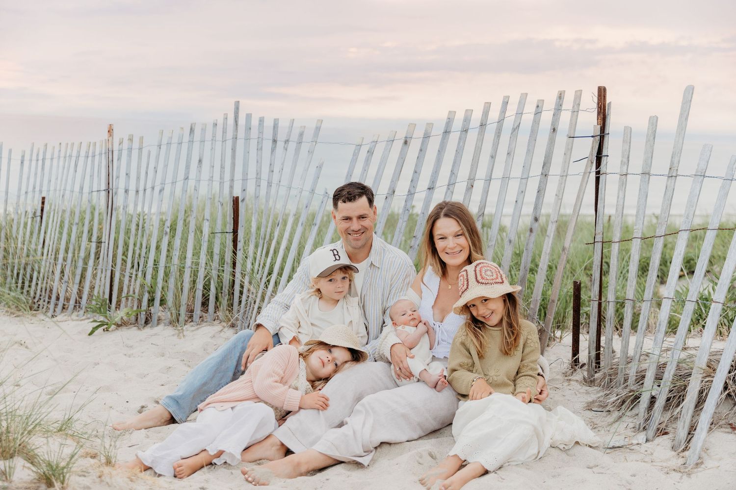 Family of six sitting together on sand dunes by a wooden beach fence at sunset.