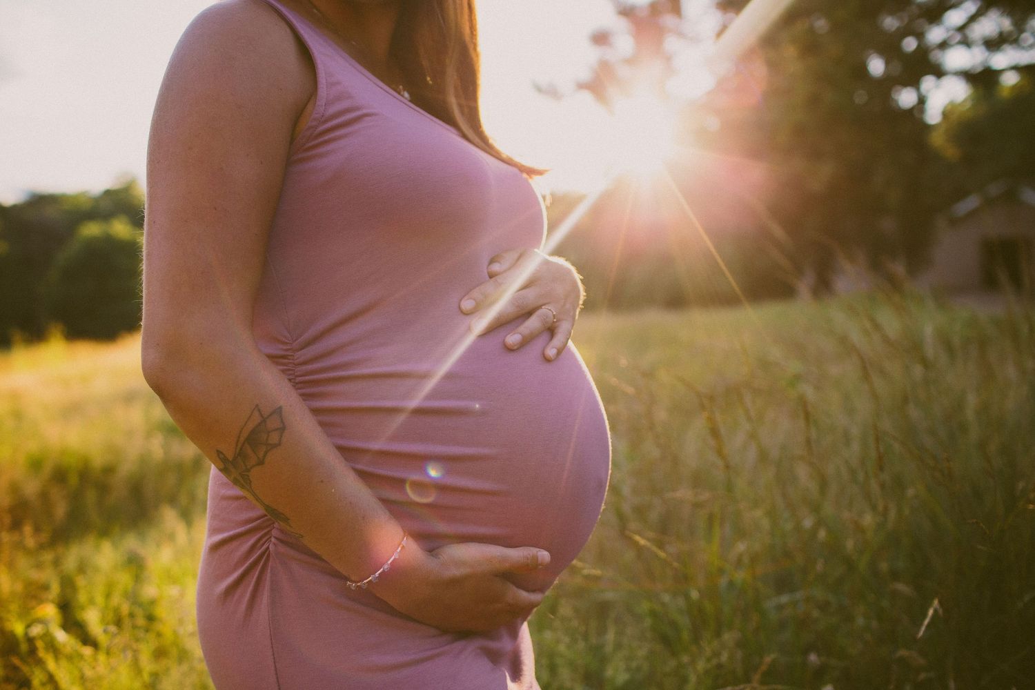 woman in a lavender maternity dress holding her pregnant belly bump while the sun shines through a field behind her