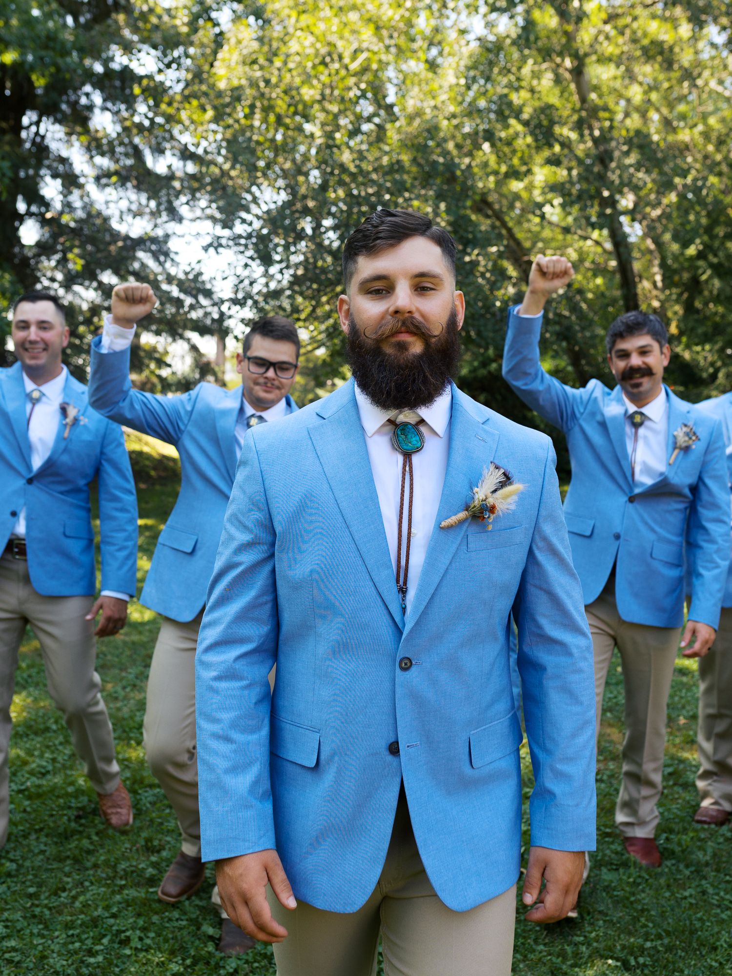 man in light blue suit jacket and handlebar mustache walks towards the camera while his groomsmen cheer him on