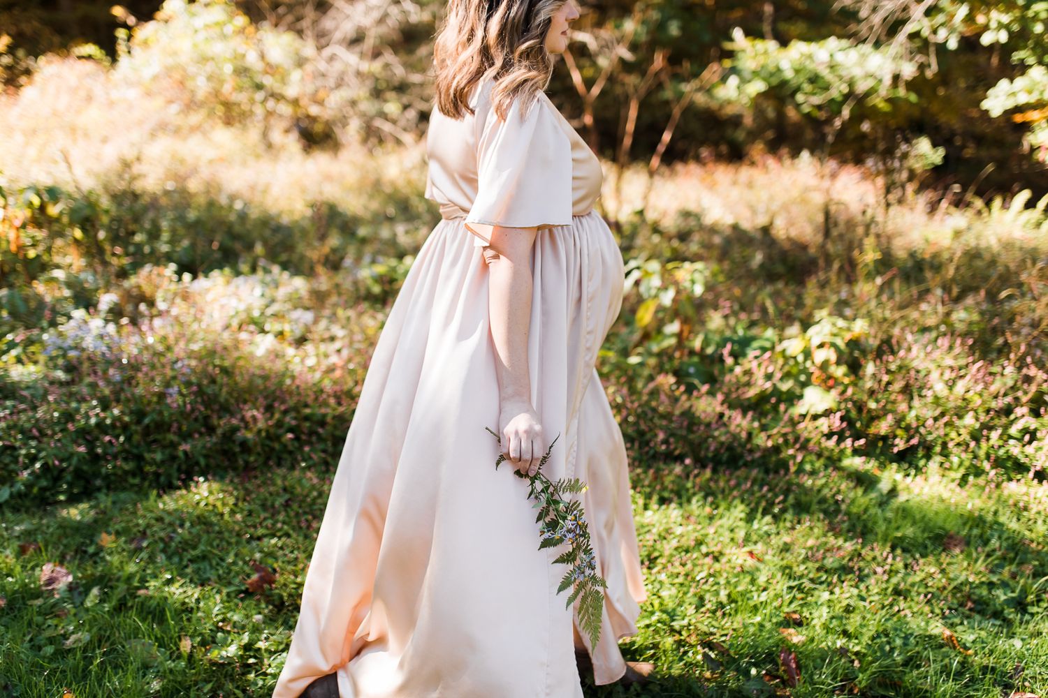 Mom walking in a silk dress with a belly bump in the Western Pennsylvania woods while holding a fern