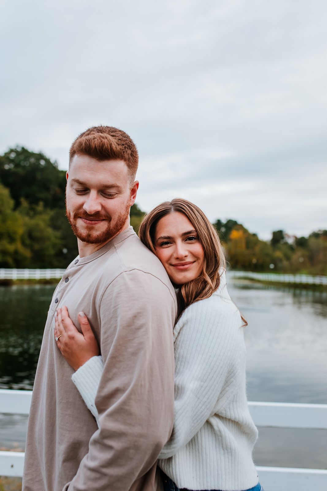 beautiful woman smirking at camera while hugging her fiance who looks over his shoulder at her with pond behind them