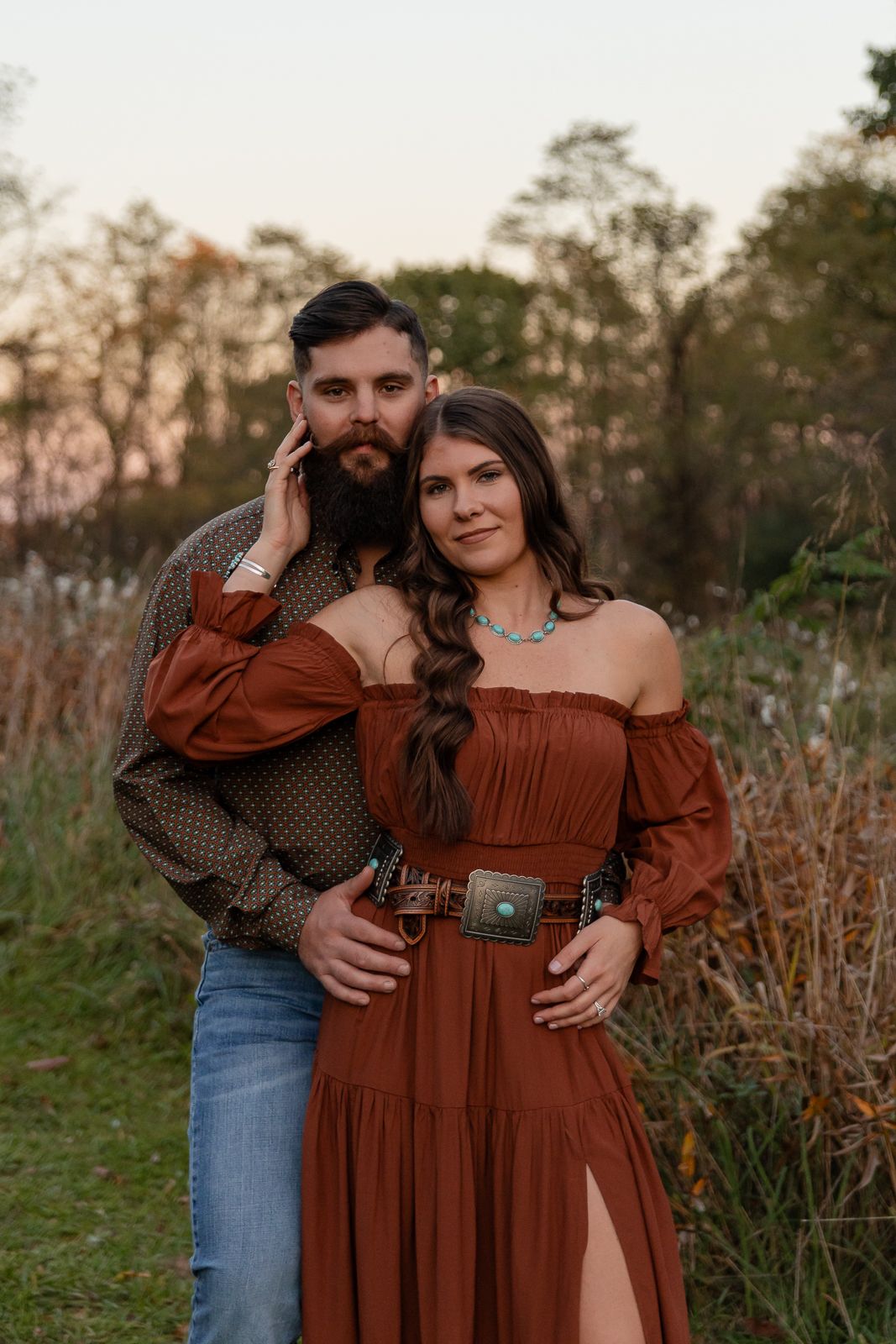 woman in rust dress being held by her fiance looking at the camera while standing in a field