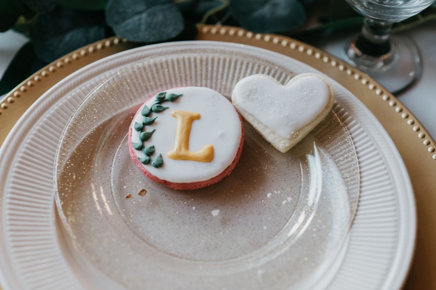 round wedding cookie with gold L and greenery on a plate with a white icing heart cookie