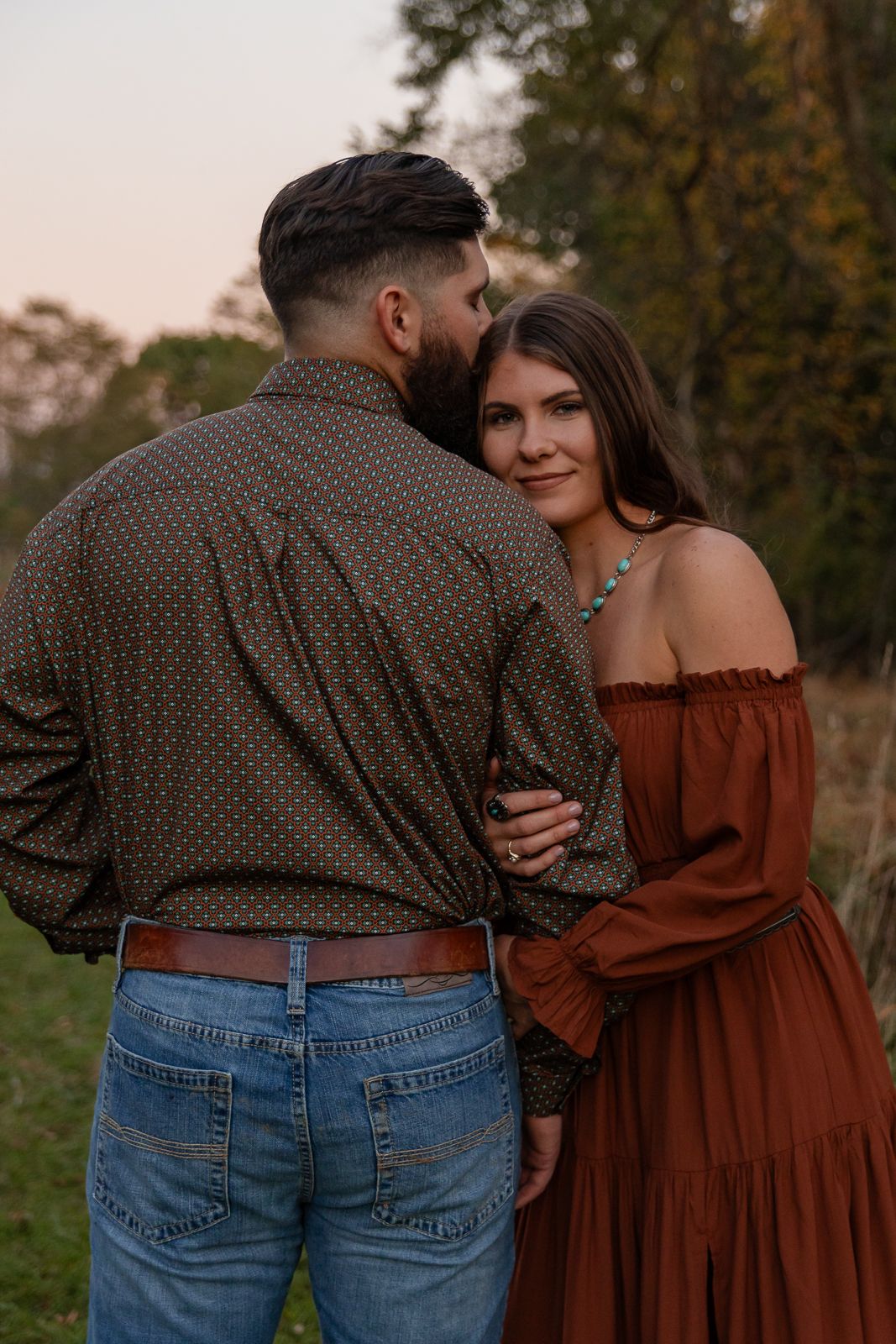 woman in rust dress holds on to man and looks at the camera while he leans over and gives her a ksis on the forehead