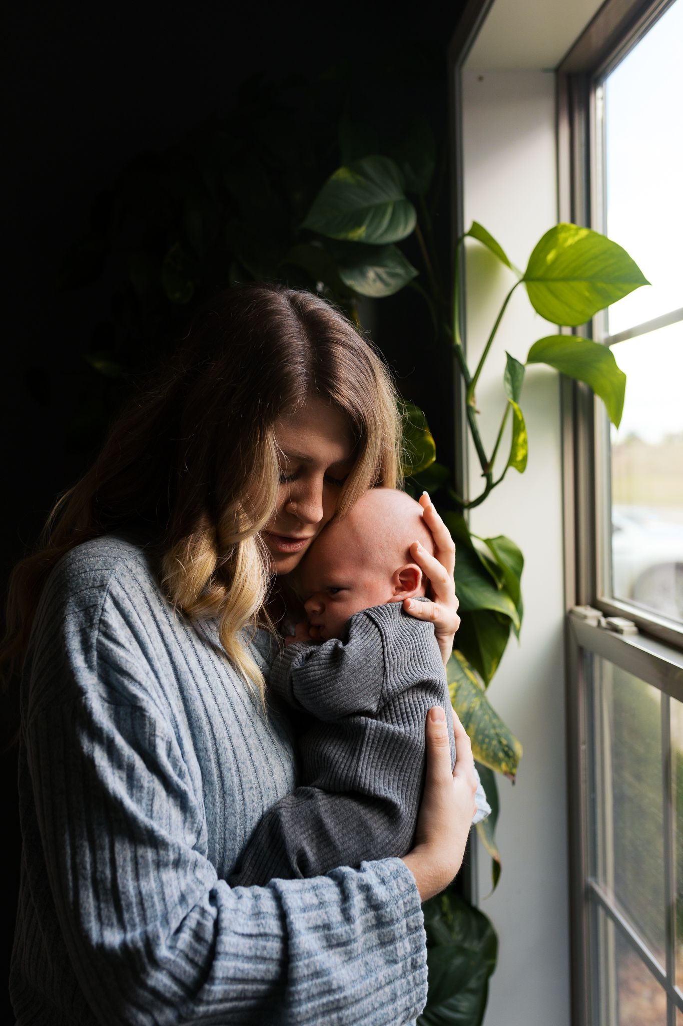 mom holding newborn baby close to her while she rocks him near a window during an in home newborn photography session
