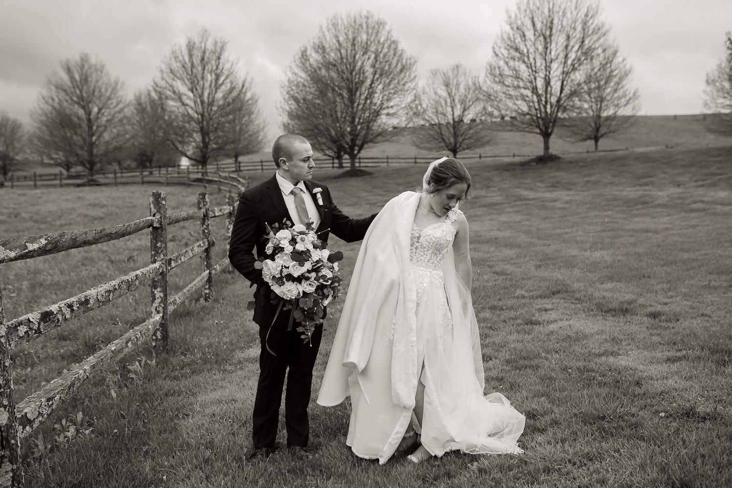 black and white image of groom holding flower bouquet while bride removes outer layer of her outfit