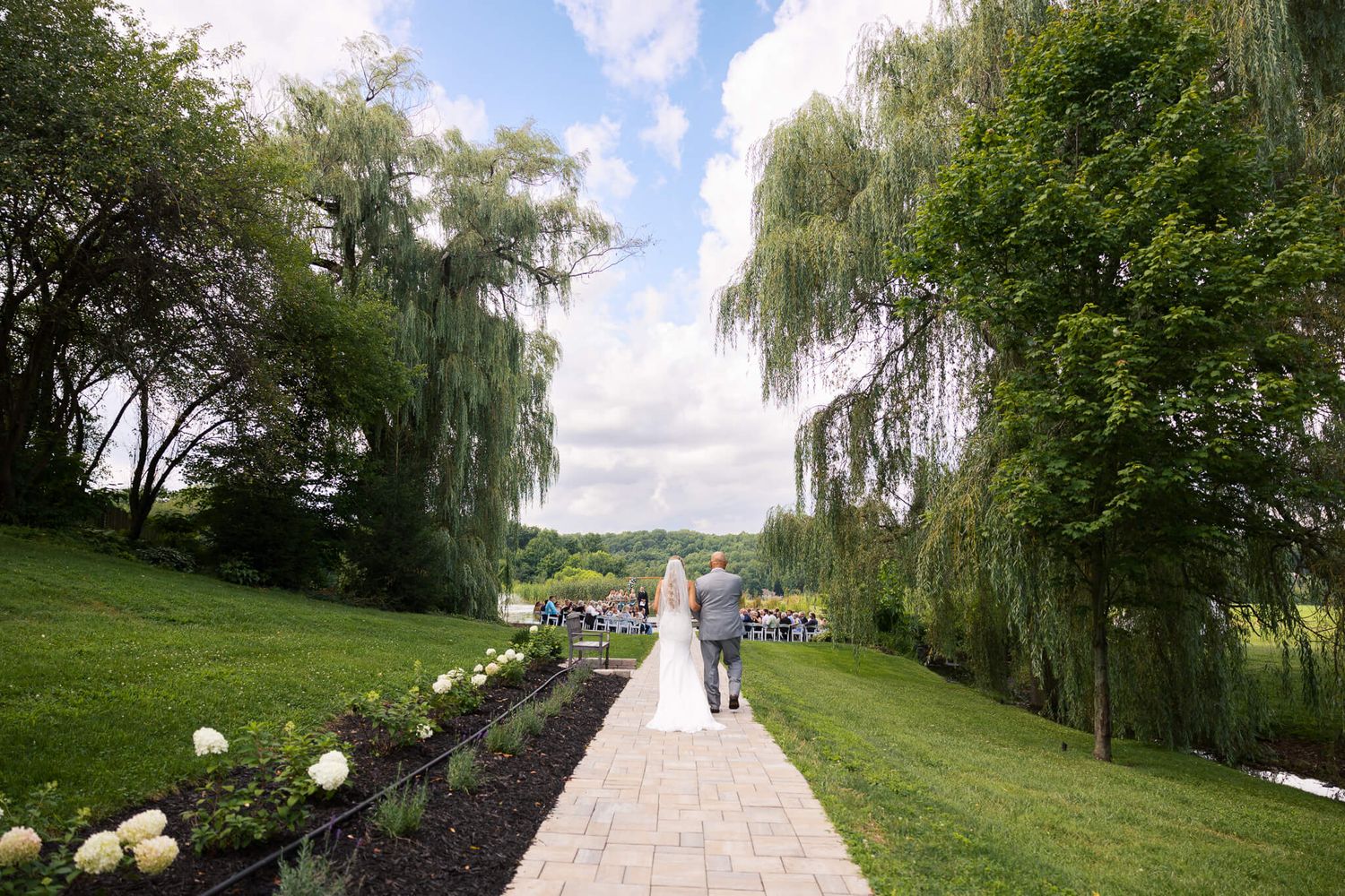 stunning image of a bride and her father walking down an outdoor brick wedding aisle surrounded by blue skies and green willow trees