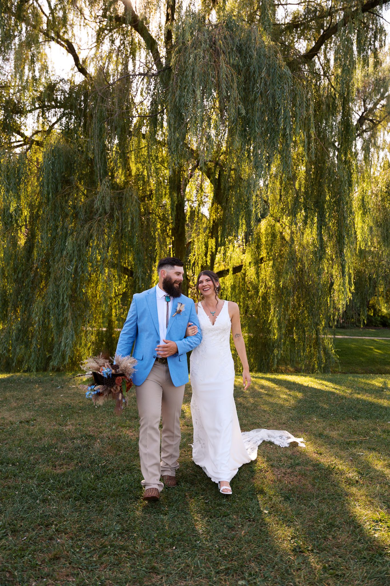 man in blue suit jacket and tan pants links arms with beautiful woman in white lace wedding gown walking beneath a willow tree in western PA