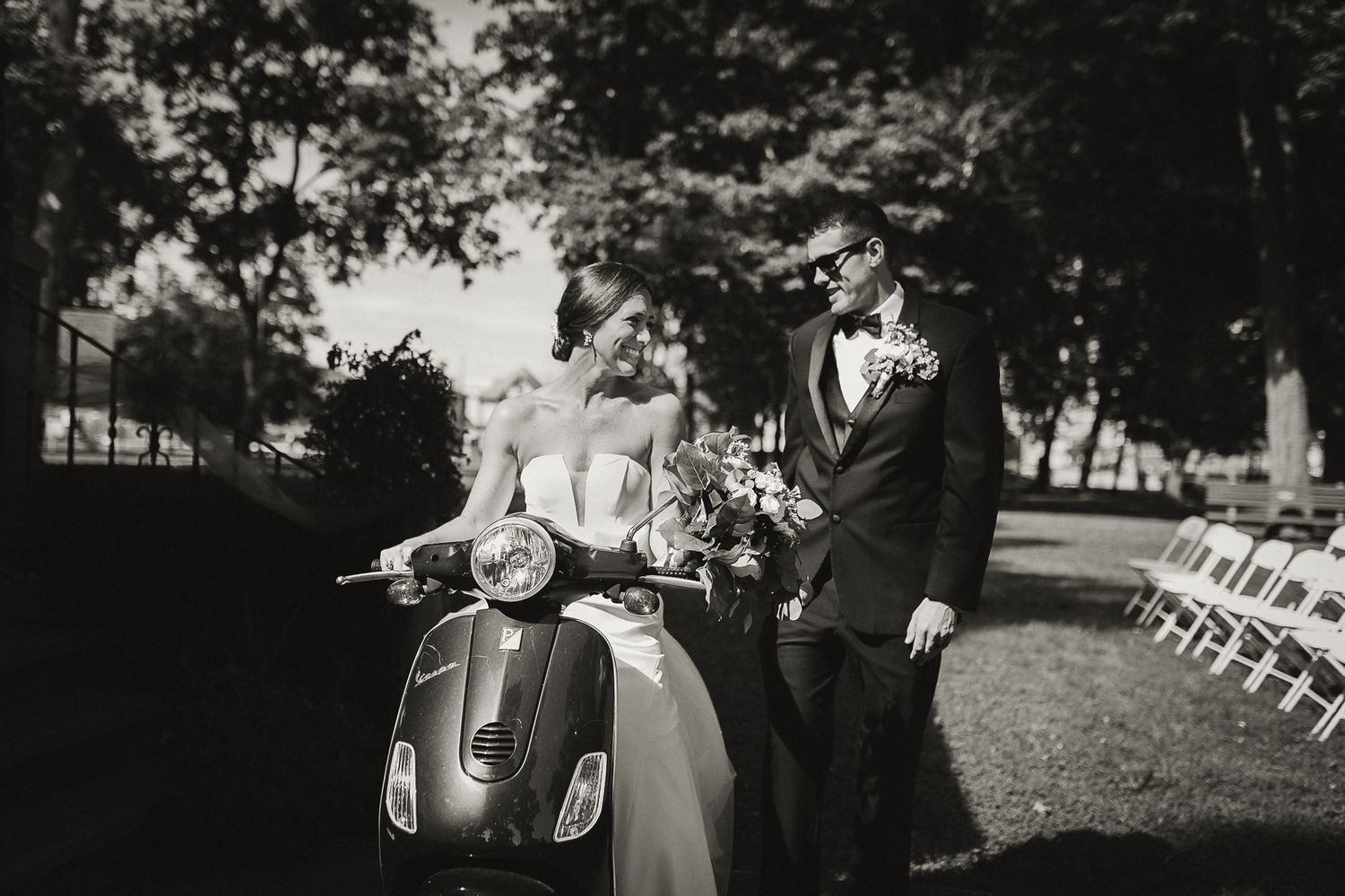 Bride sitting on vespa looking happily at her husband in a tux and sunglasses standing beside her