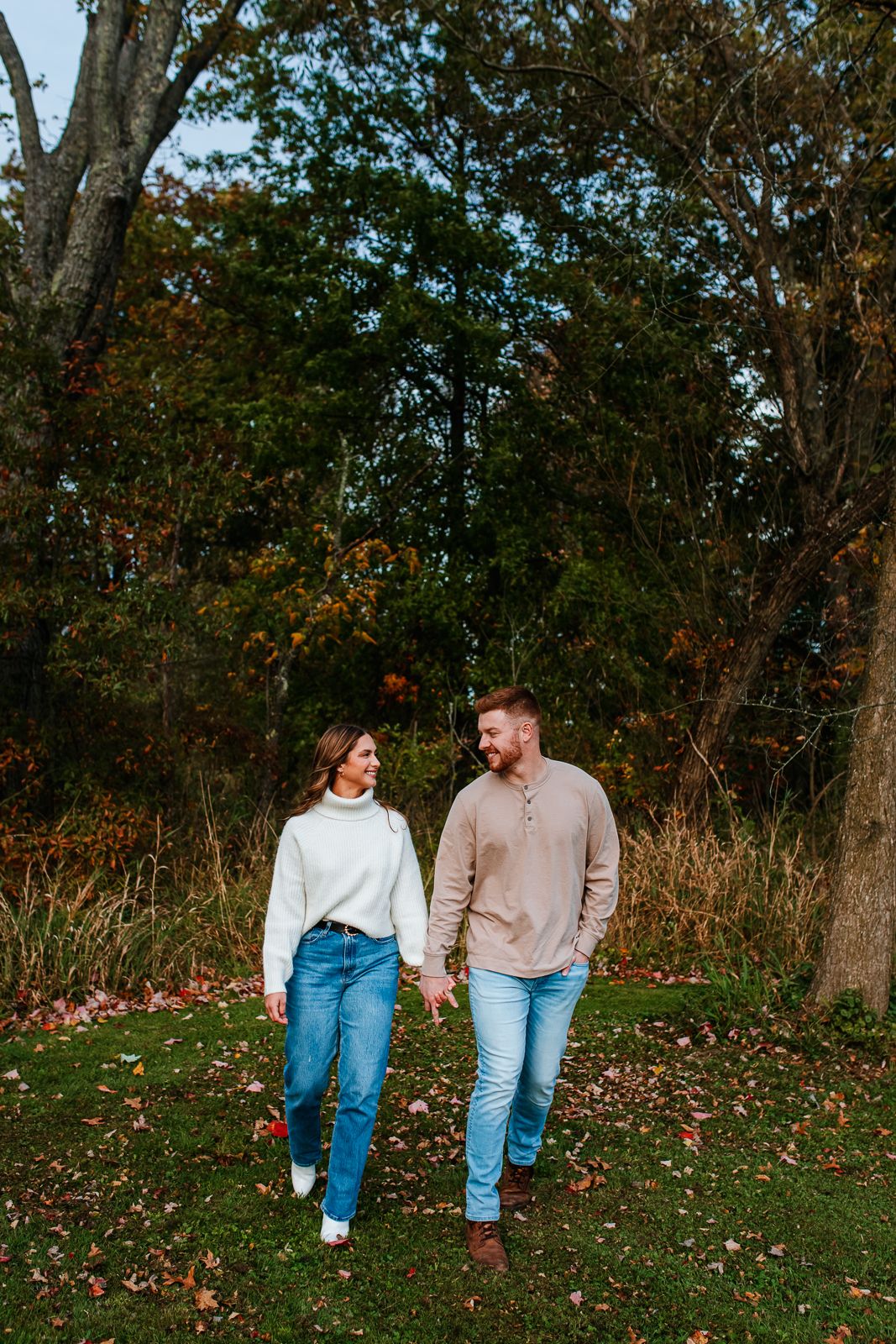 man and woman in comfy sweaters holding hands and walking towards camera during playful engagement posing