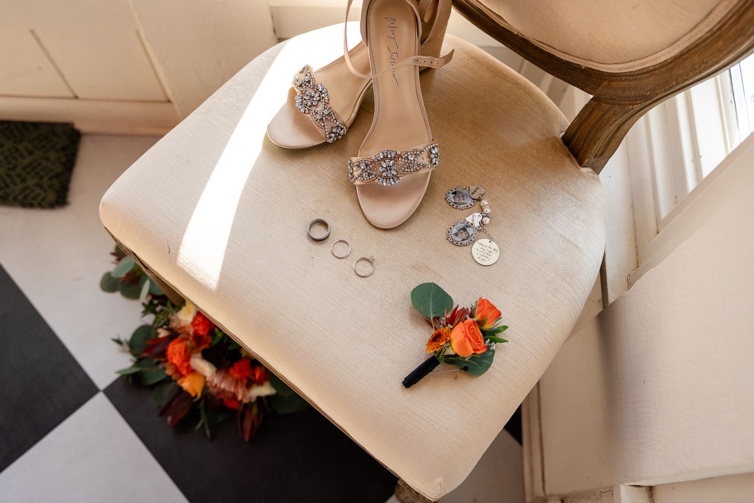 wedding shoes, rings, bouquet charms and flower sitting on a luxury white chair