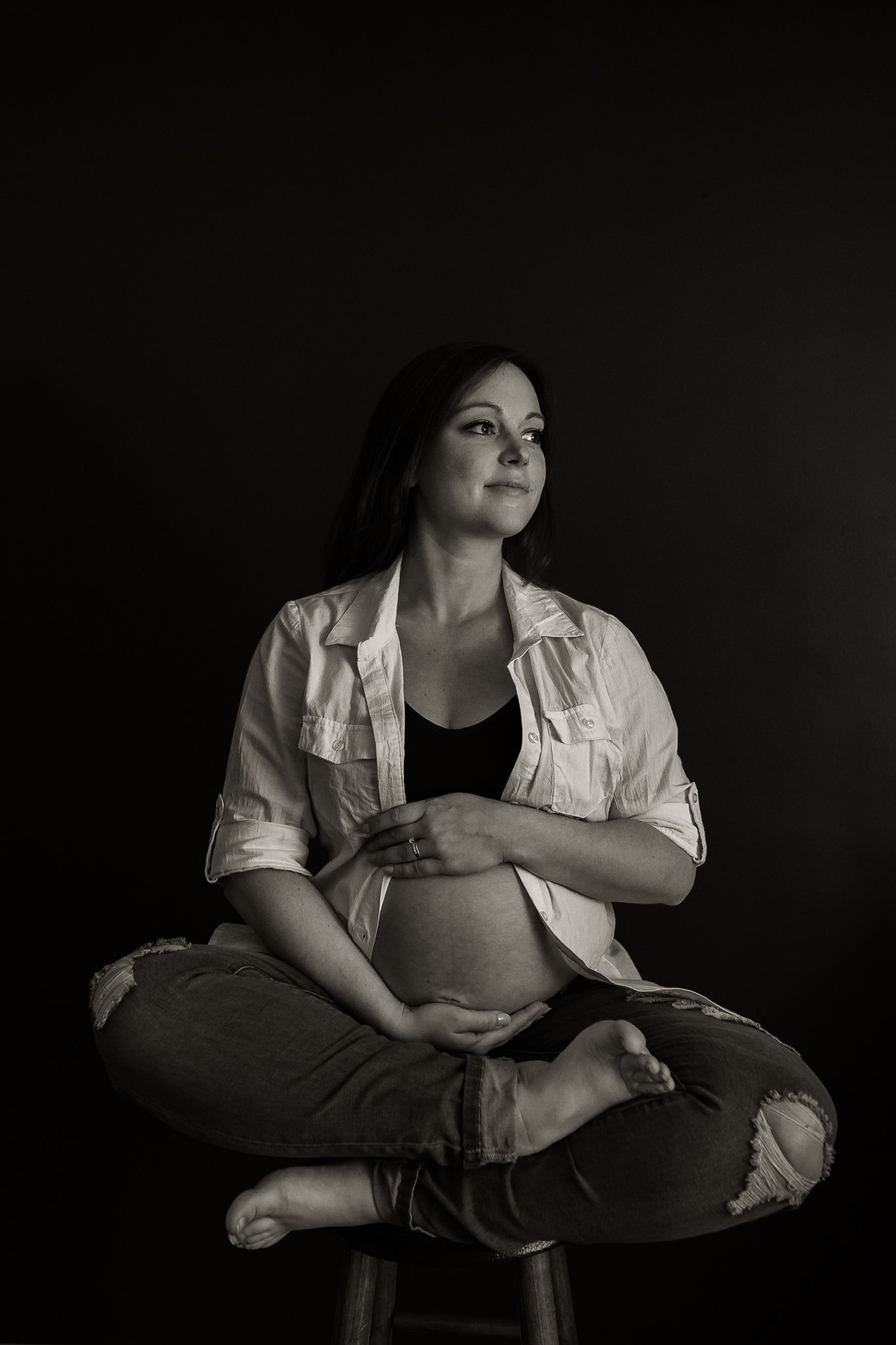 mom to be sitting cross legged on a stool holding her pregnant belly with both hands in black and white