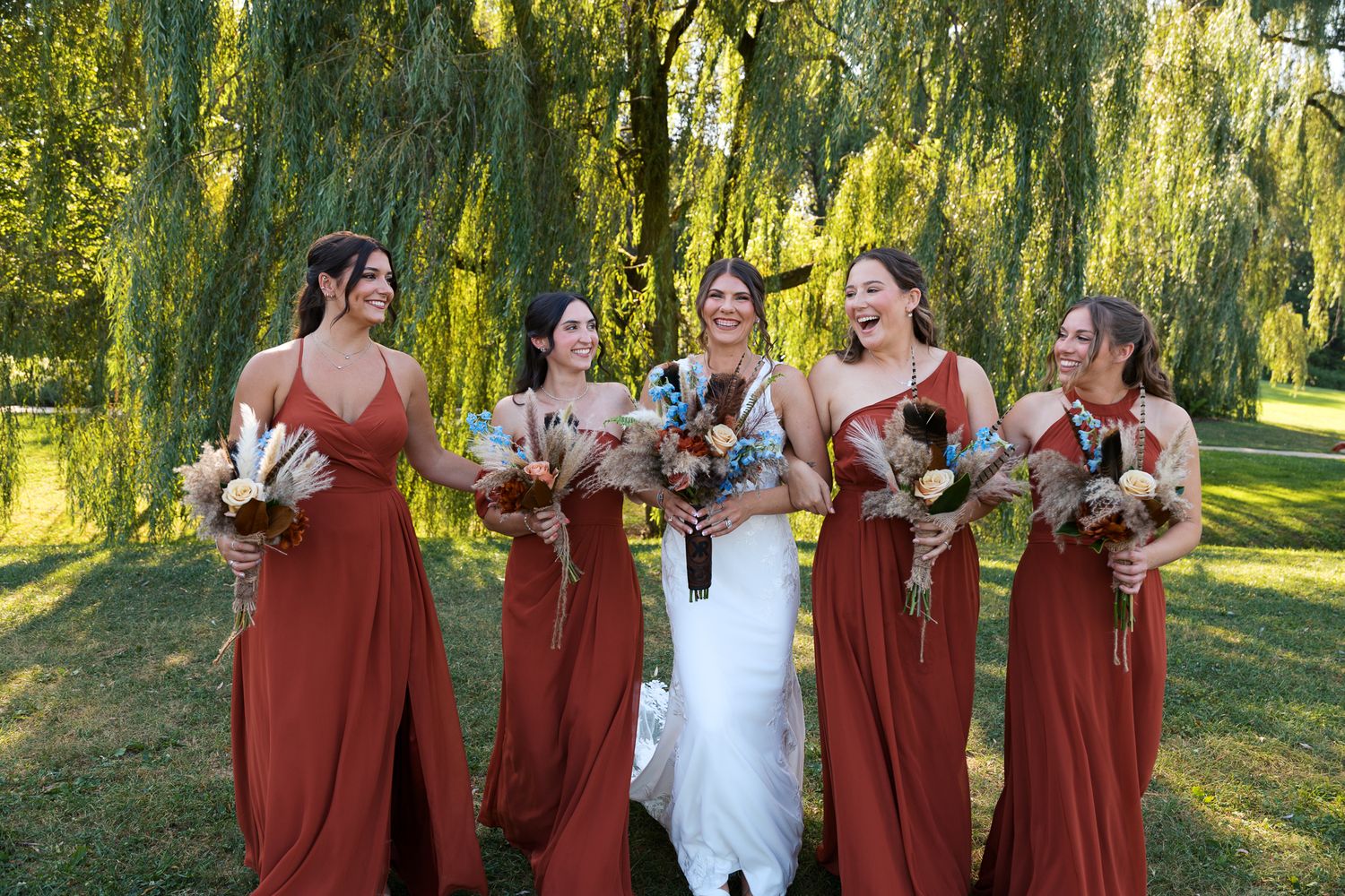bridal party in rust dresses with turkey feather flower bouquets linked arms with bride in white lace wedding gown laughing and walking