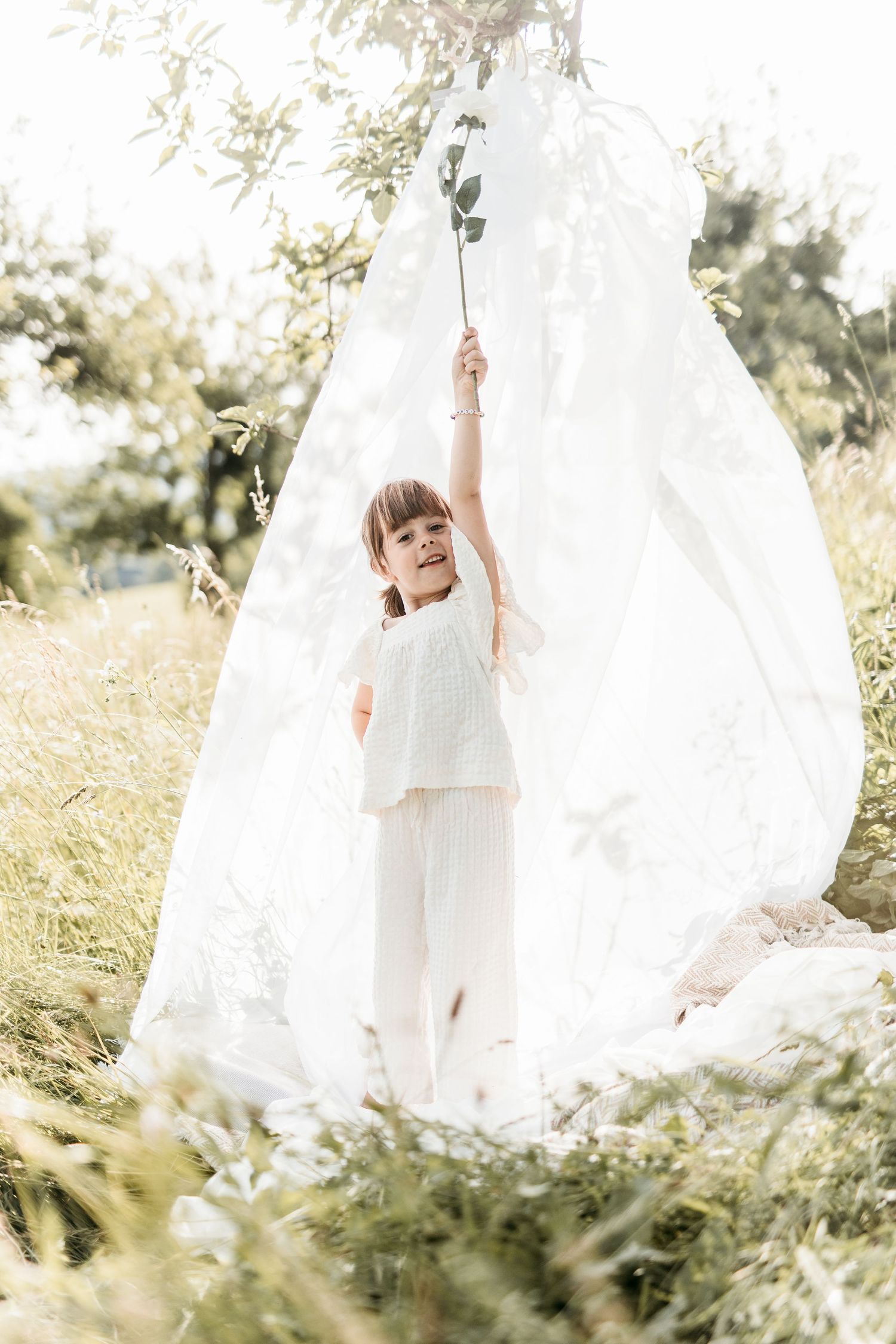Little girl in white clothes reaching up holding a flower stem inside a white fabric teepee outdoors.