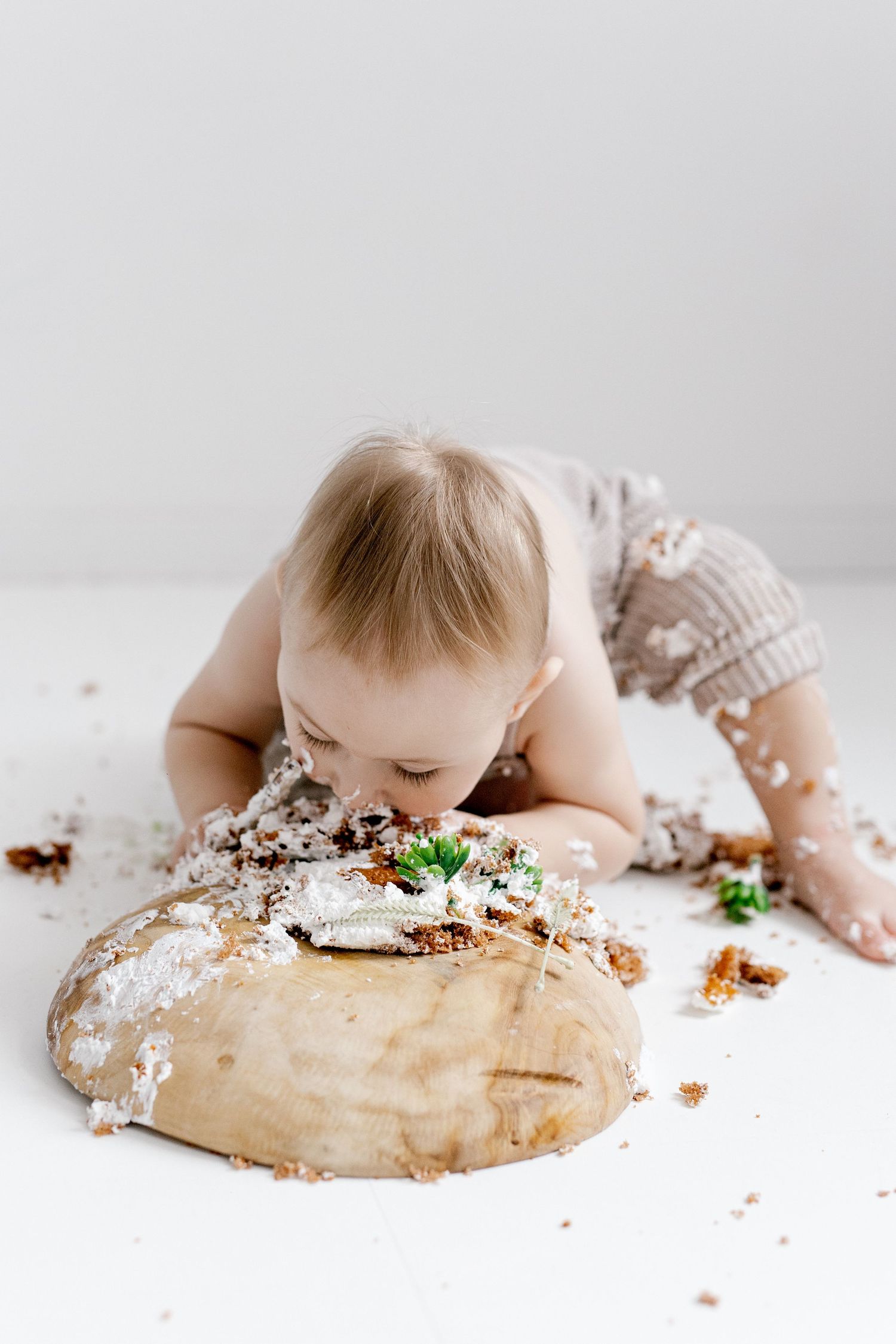 Baby in knit romper face-down eating smashed first birthday cake on wooden board.