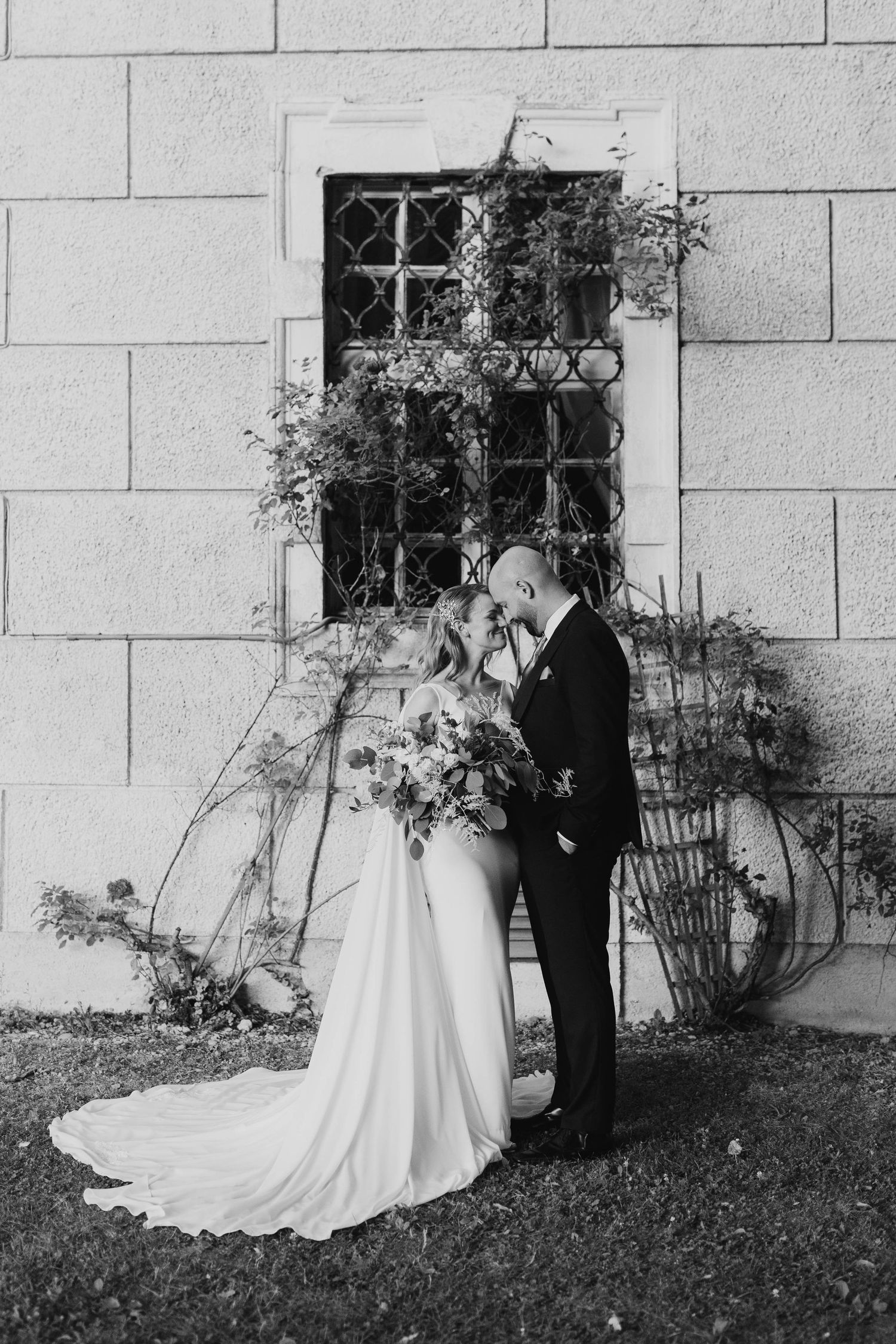 Black and white photo of bride and groom kissing before a stone wall with vintage window and climbing vines.