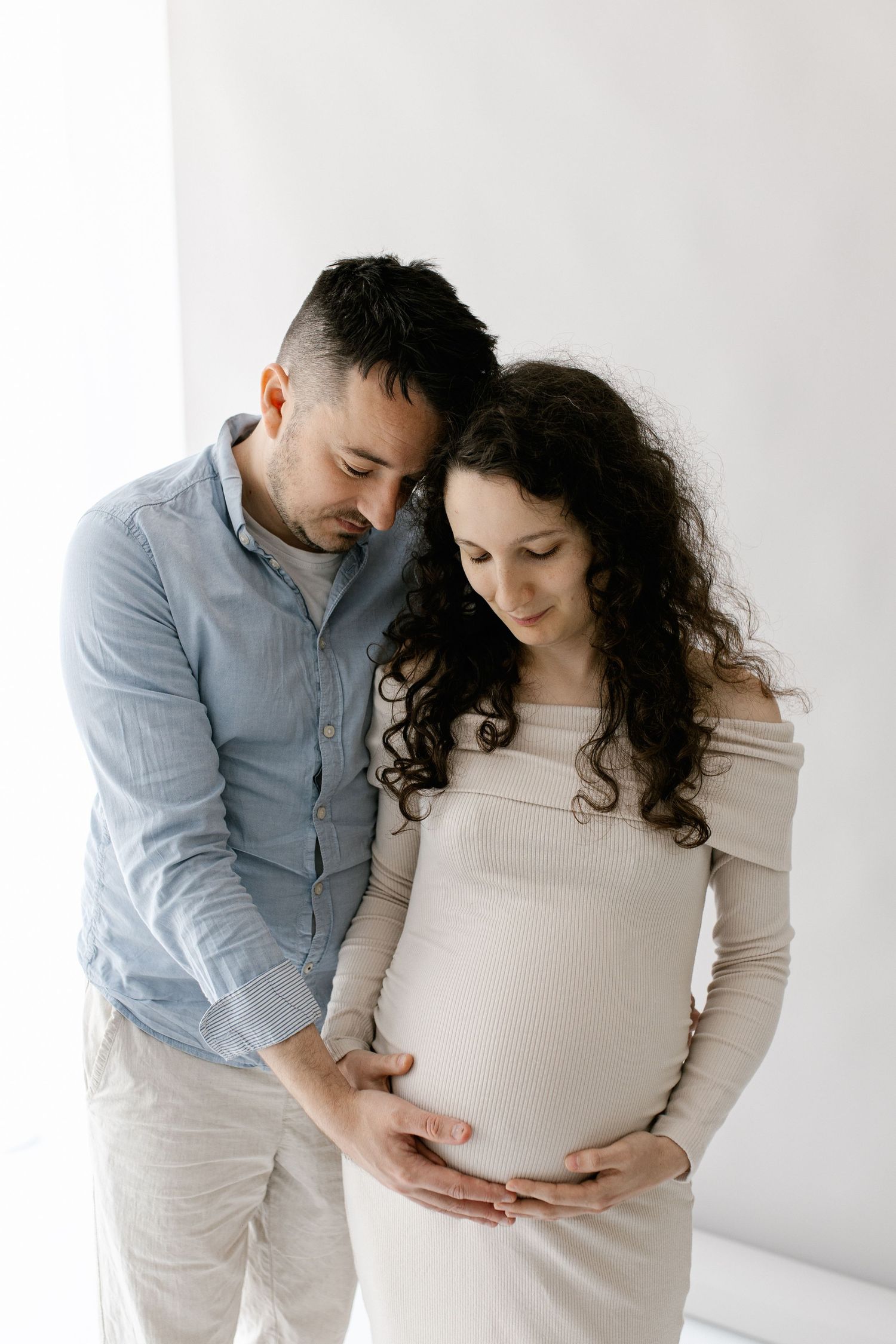 Couple embracing with man holding pregnant woman's belly in bright white studio setting.