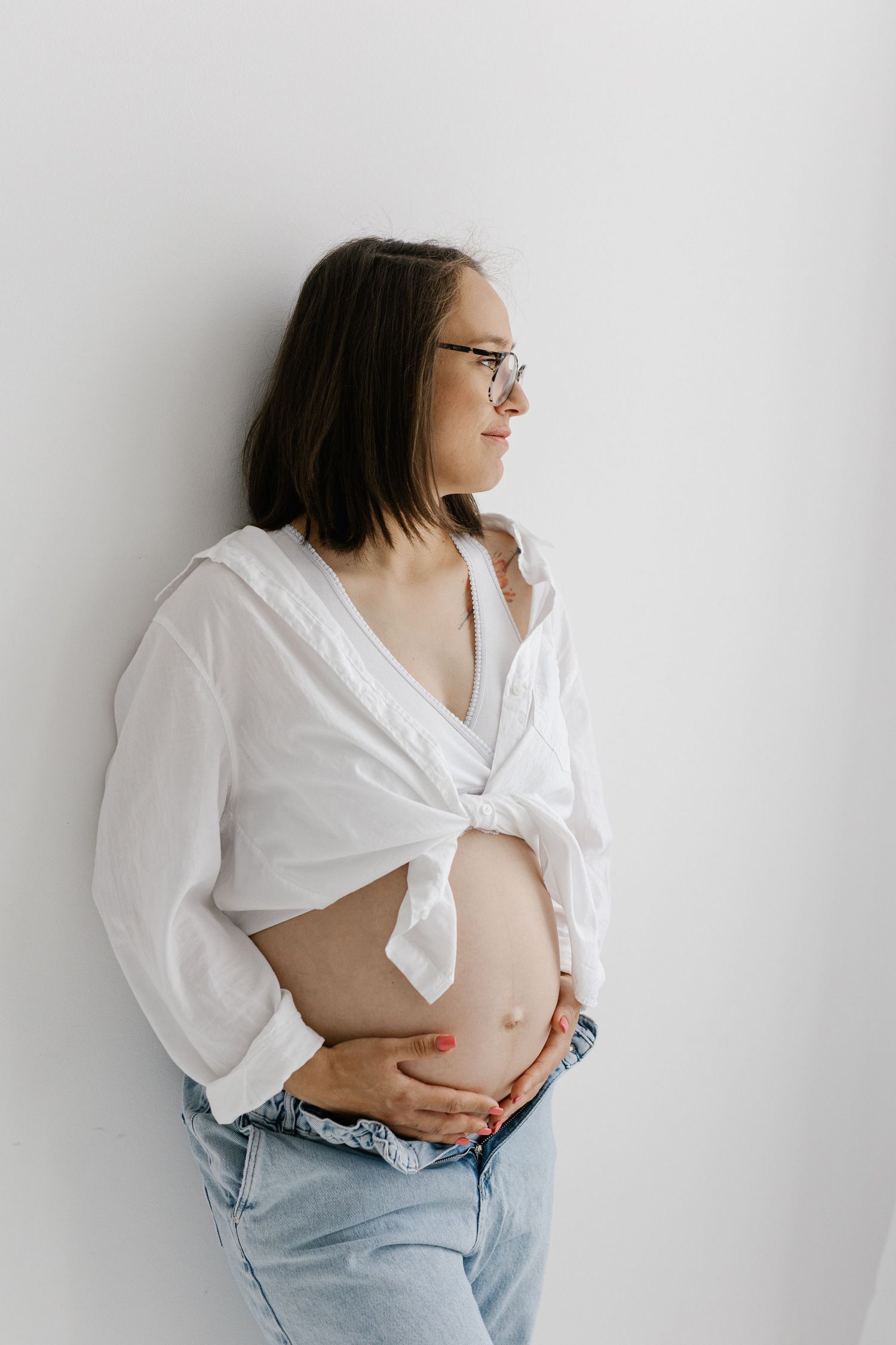 Pregnant woman wearing glasses and white tied shirt holding her baby bump while looking sideways.