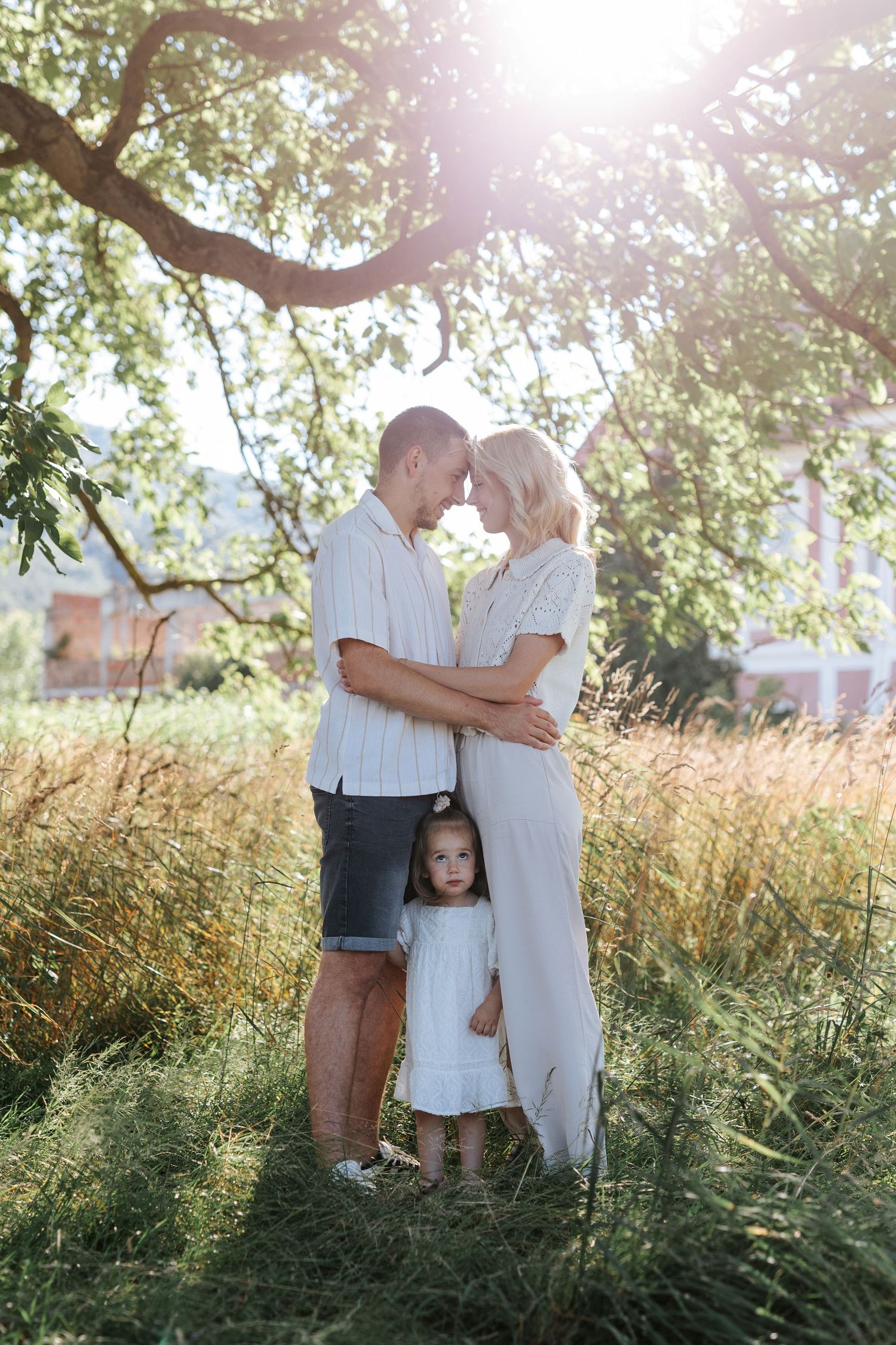 Couple touching foreheads tenderly in sunlit meadow while young daughter in white dress stands between them.