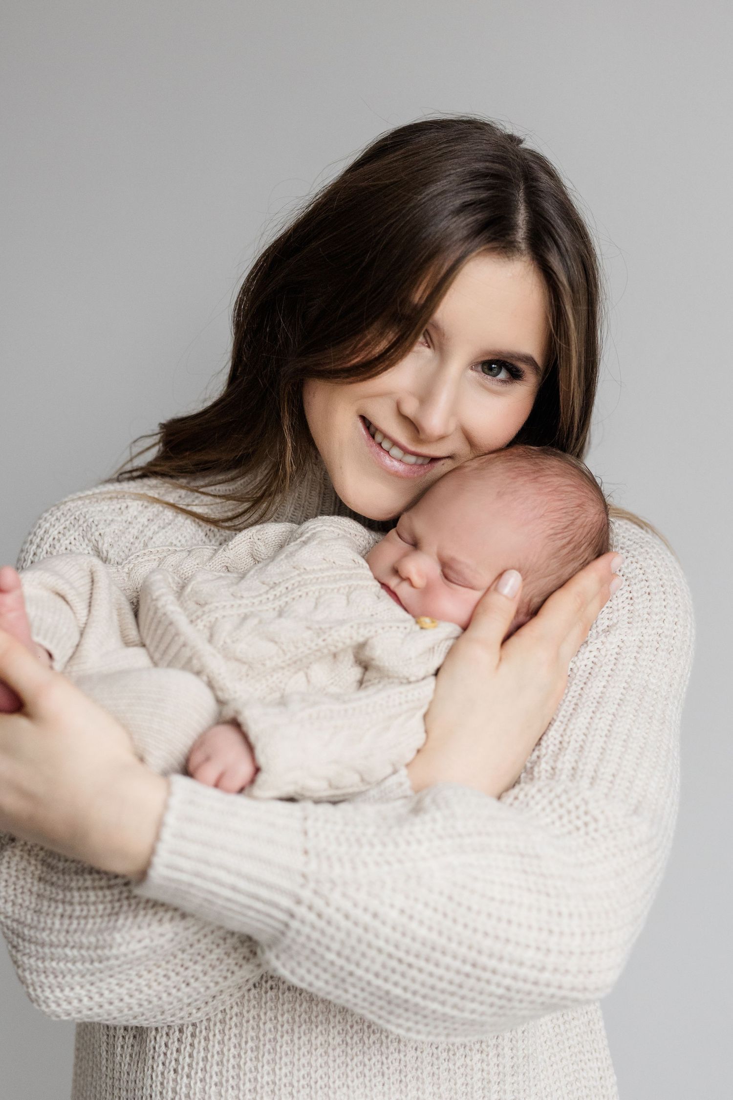 Happy brunette mother in cream knit sweater cuddles sleeping newborn baby against her chest in studio portrait.